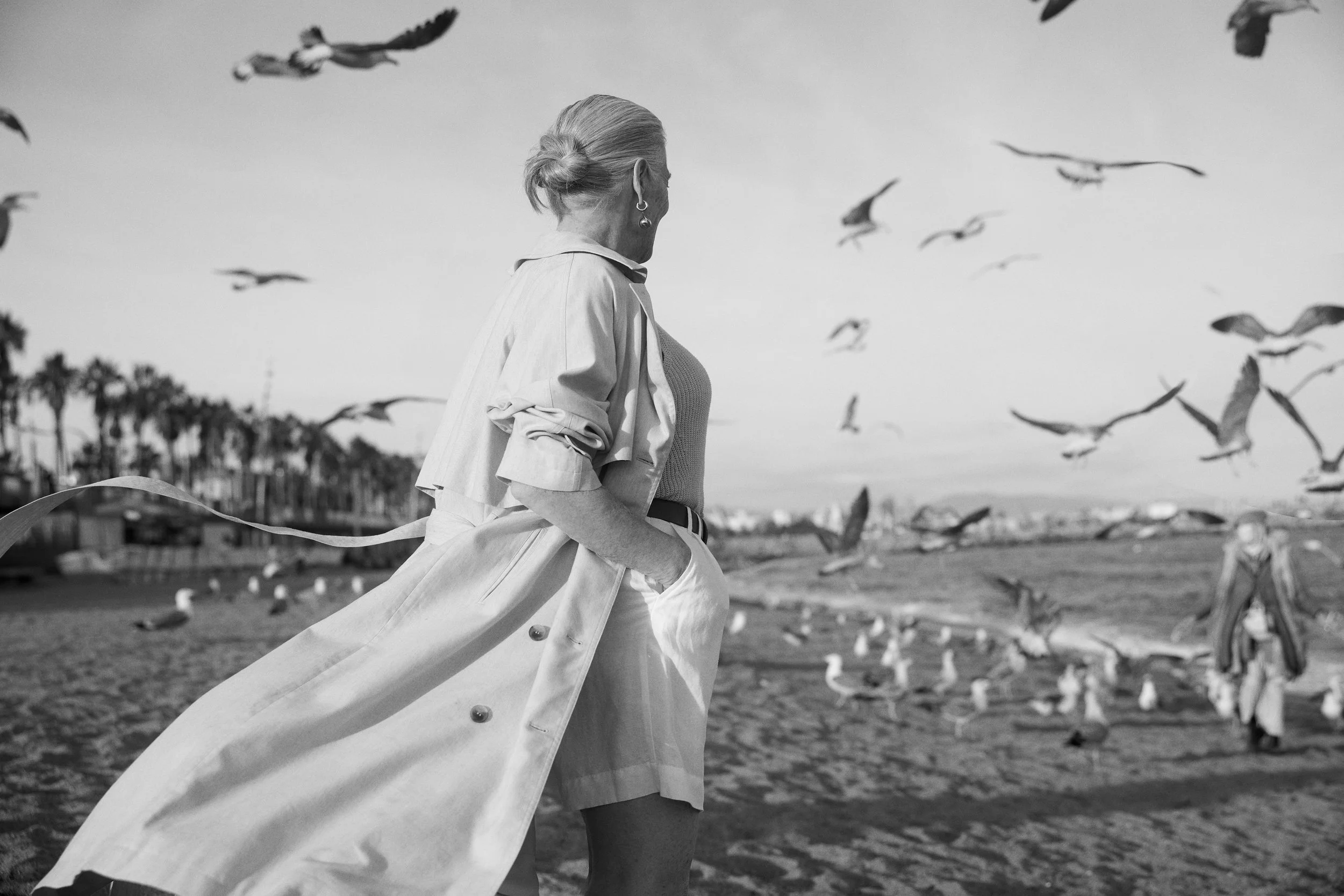 A woman with short hair wearing a light-colored coat over a dress, standing on a beach, watching seagulls fly in the sky, palm trees in the distance, and a person near seagulls in the background, all in black and white.