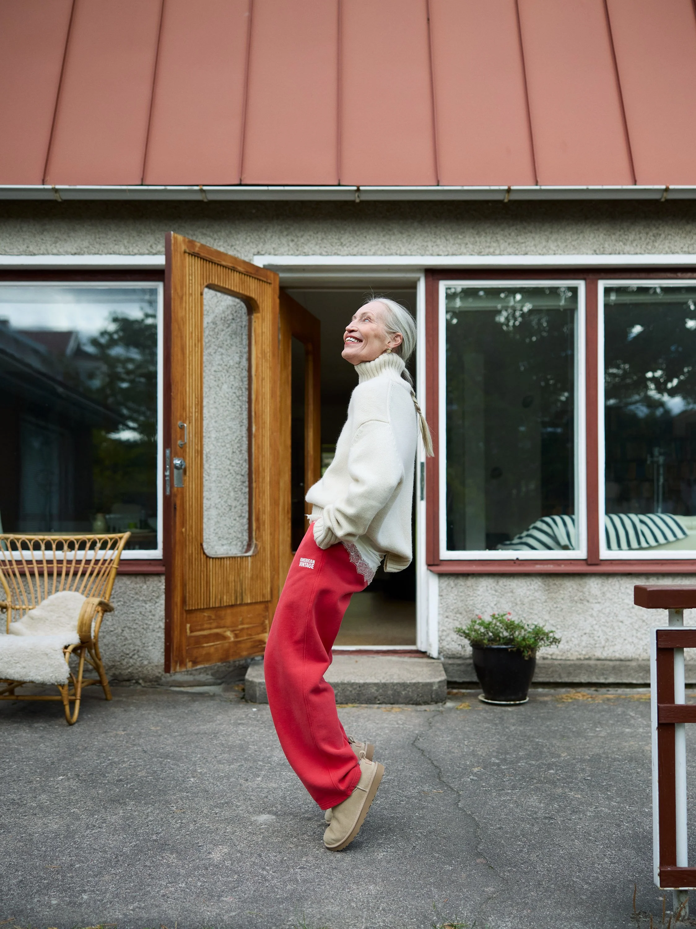 An elderly woman in a cream turtleneck sweater and red sweatpants standing outside a house, smiling and leaning back.