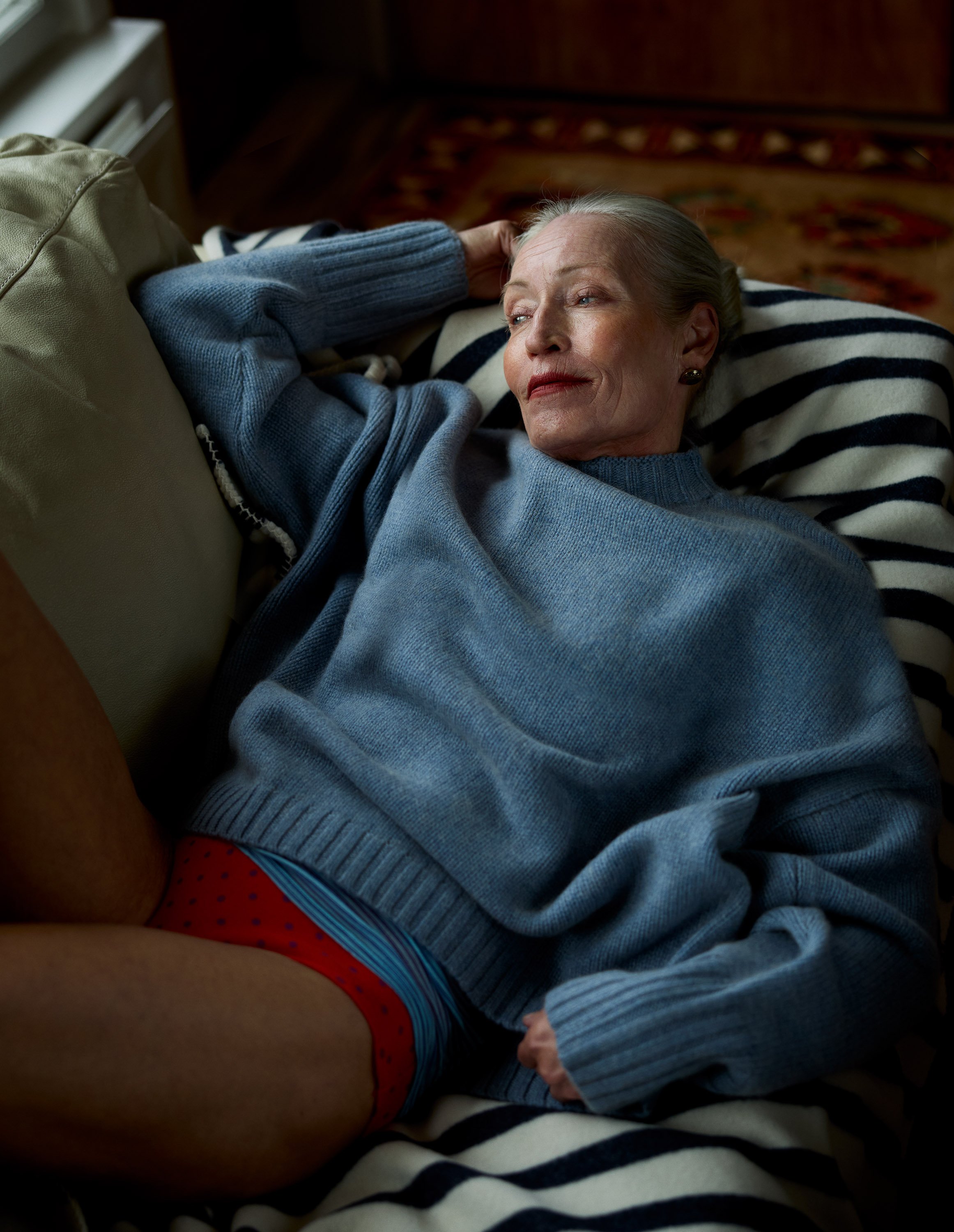 An elderly woman lying on a striped blanket, wearing a blue sweater and red polka dot underwear, with her eyes closed and a relaxed expression.