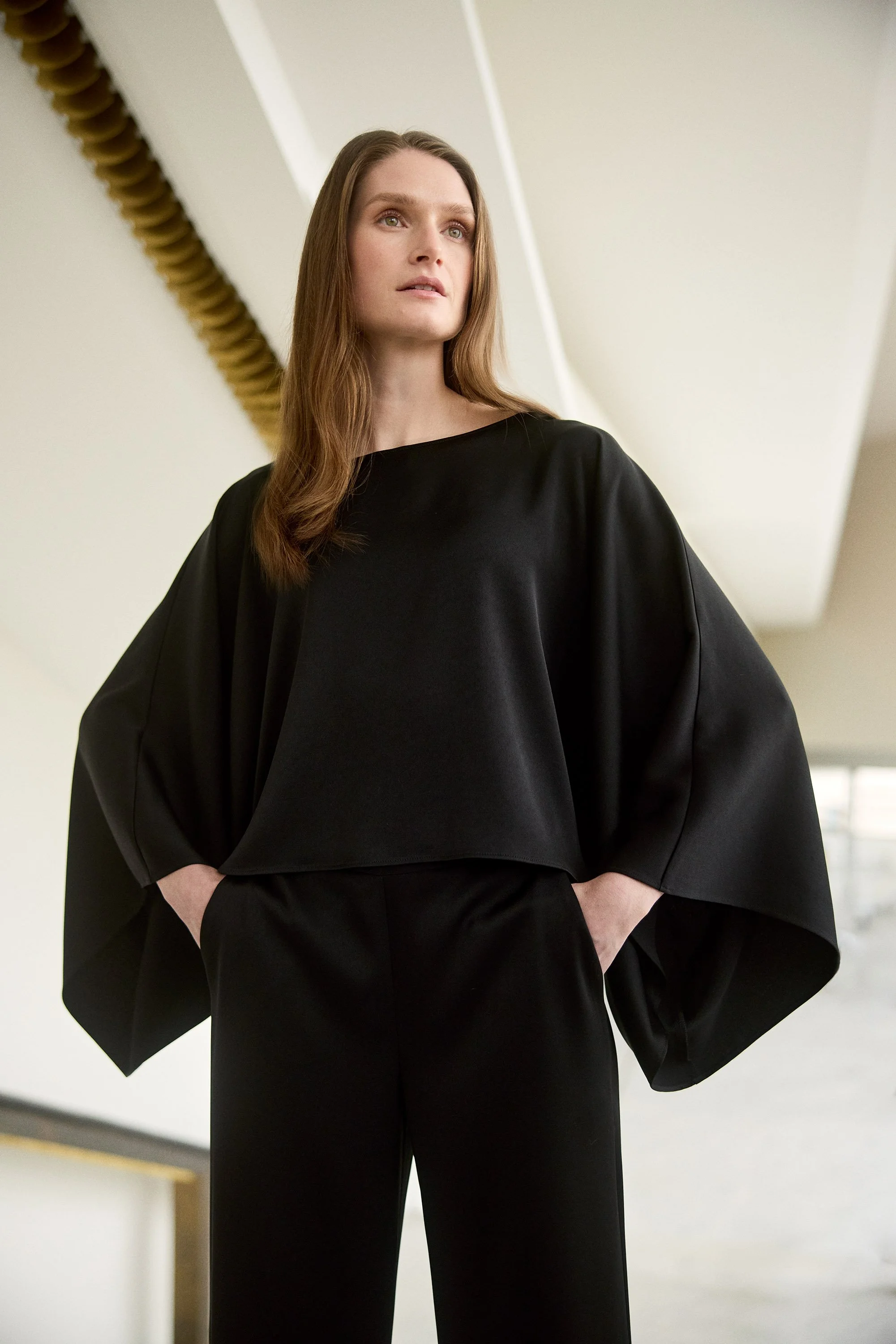 A woman with long brown hair wearing a loose black top and matching black pants, standing with her hands in her pockets, looking slightly upward, in an indoor space.