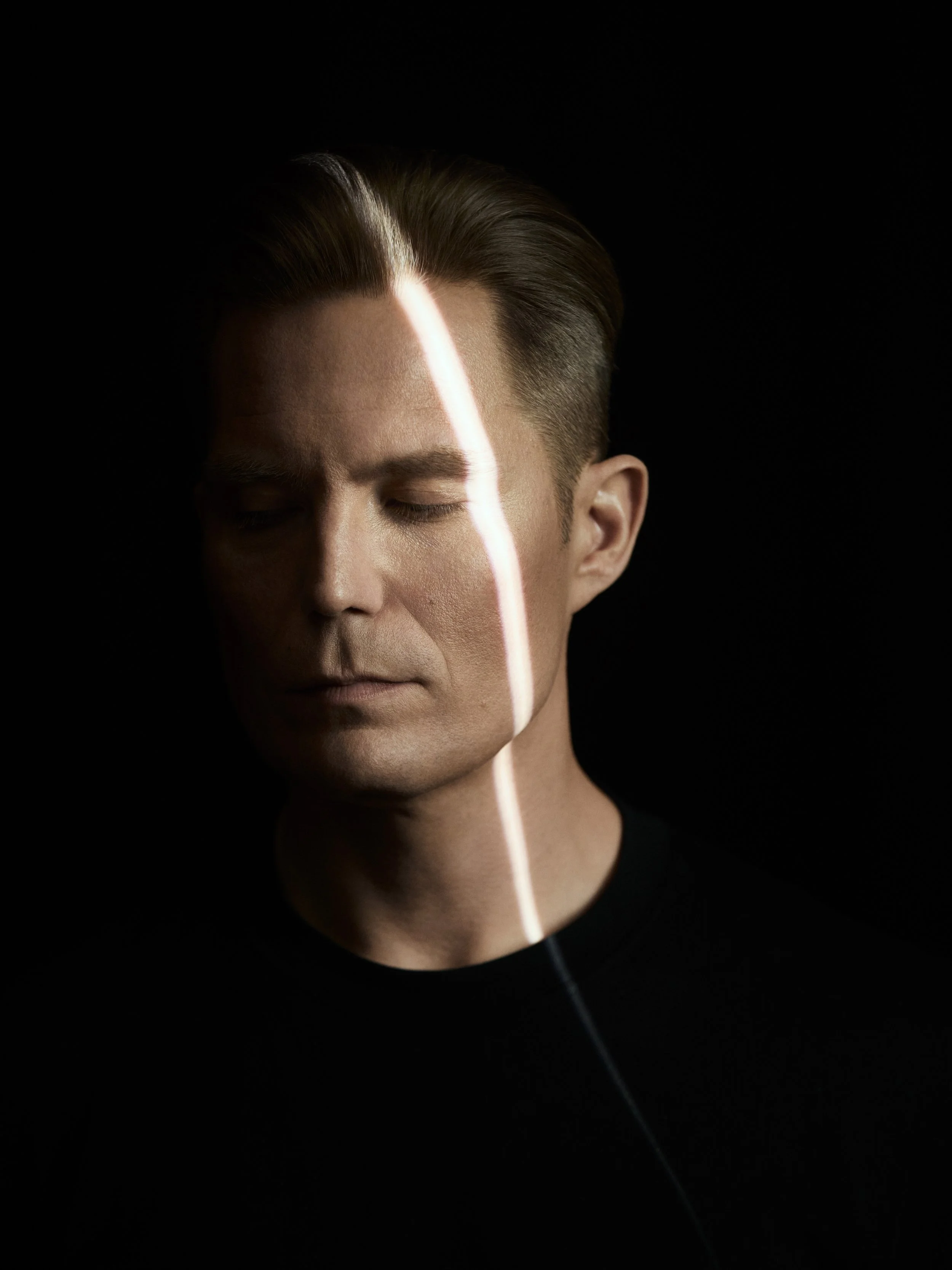A man with short, styled hair and closed eyes, with a vertical streak of light crossing his face against a dark background.