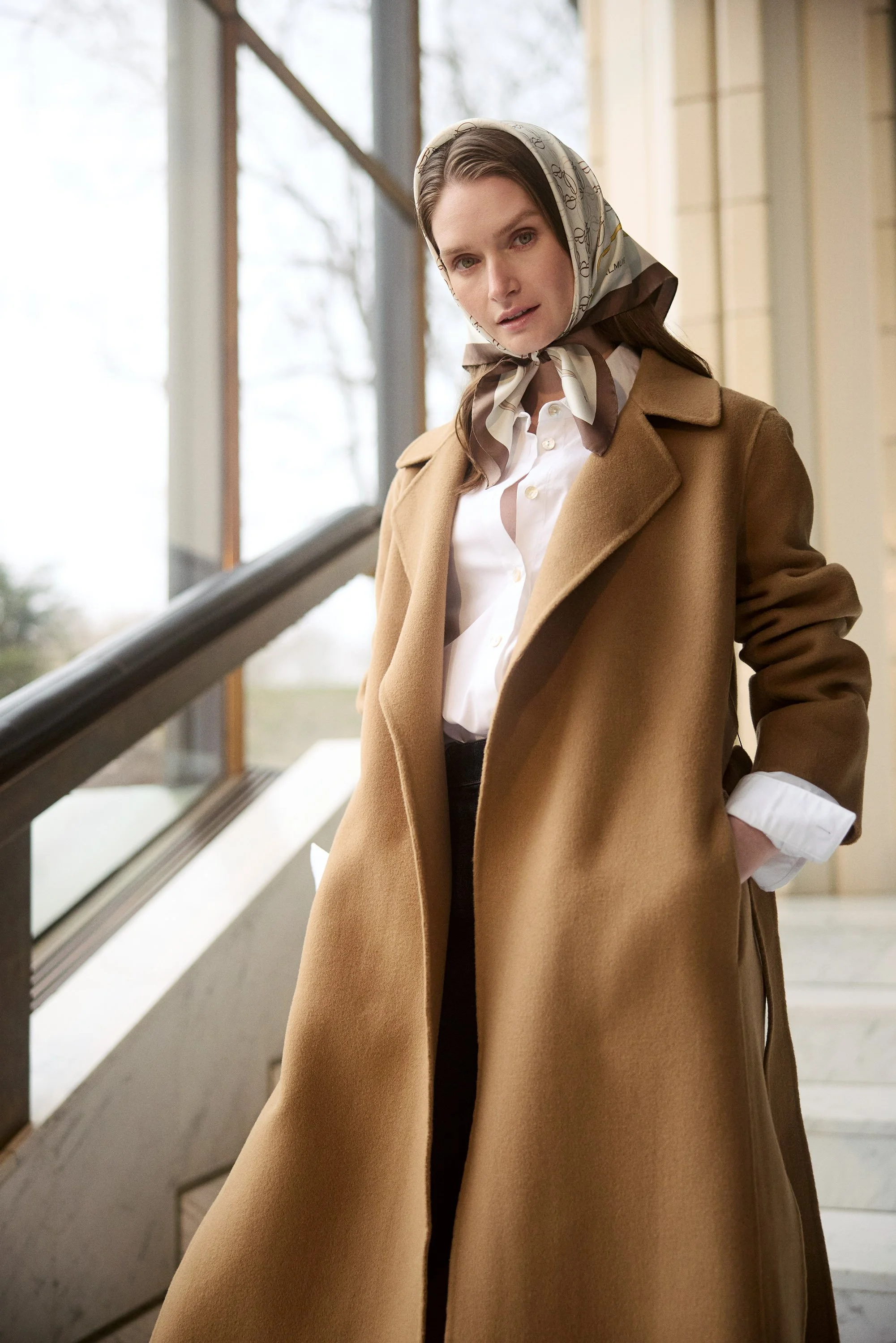 A woman with long brown hair wearing a beige coat, white shirt, and a patterned headscarf standing on stairs inside a building.