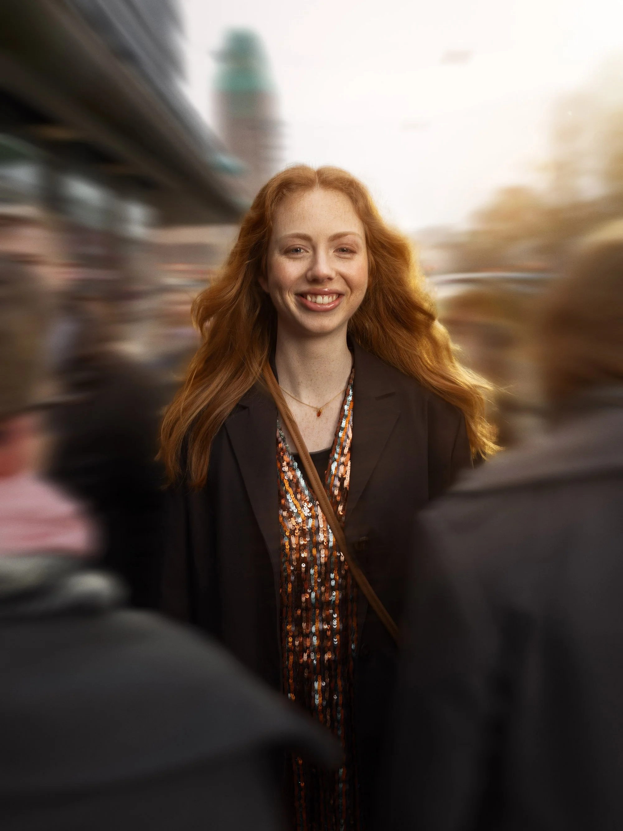 A woman with long red hair wearing a black blazer and a sequined top, smiling, in a crowd with a blurred background.