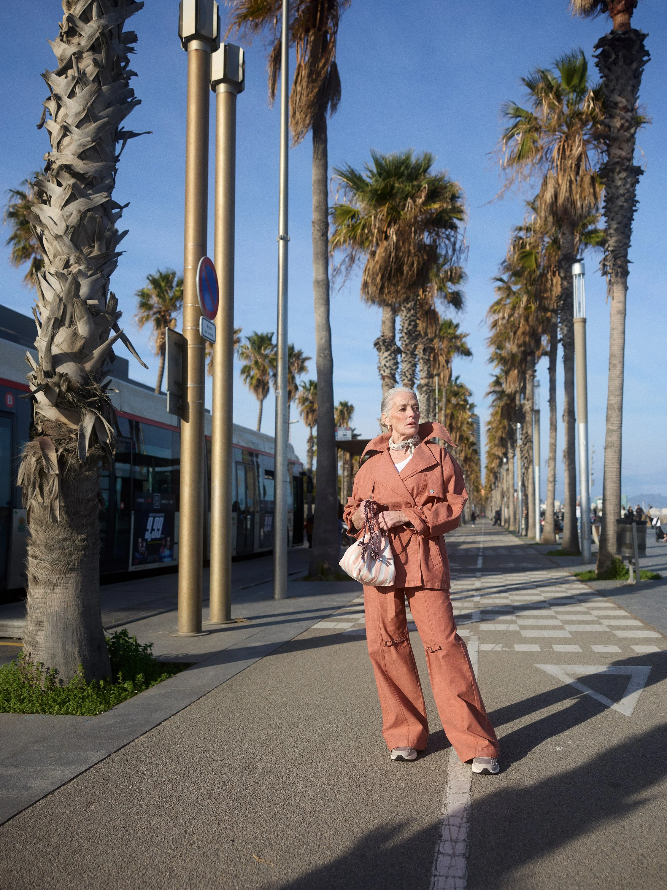 An elderly woman standing on a sidewalk lined with palm trees, wearing a salmon-colored outfit and holding a striped bag, with a bus and a row of palm trees in the background.