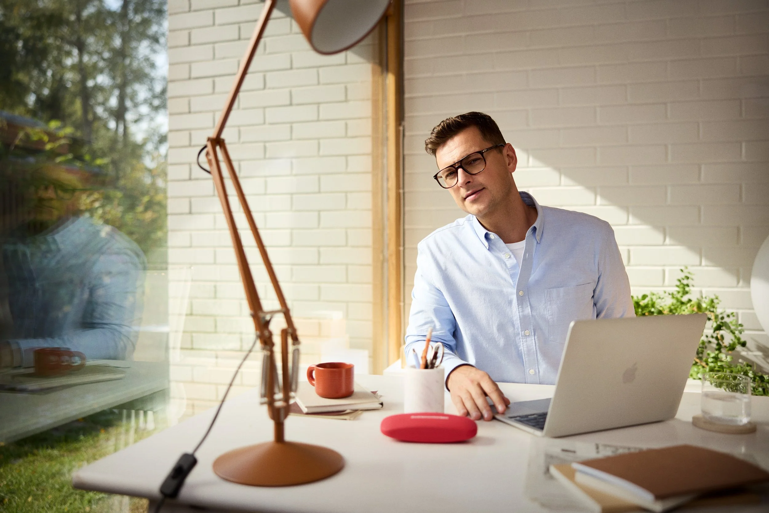 A man in glasses and a light blue shirt working on a laptop at a white desk with a desk lamp, cup, notebook, and pens, in a room with white brick walls and a window with greenery outside.