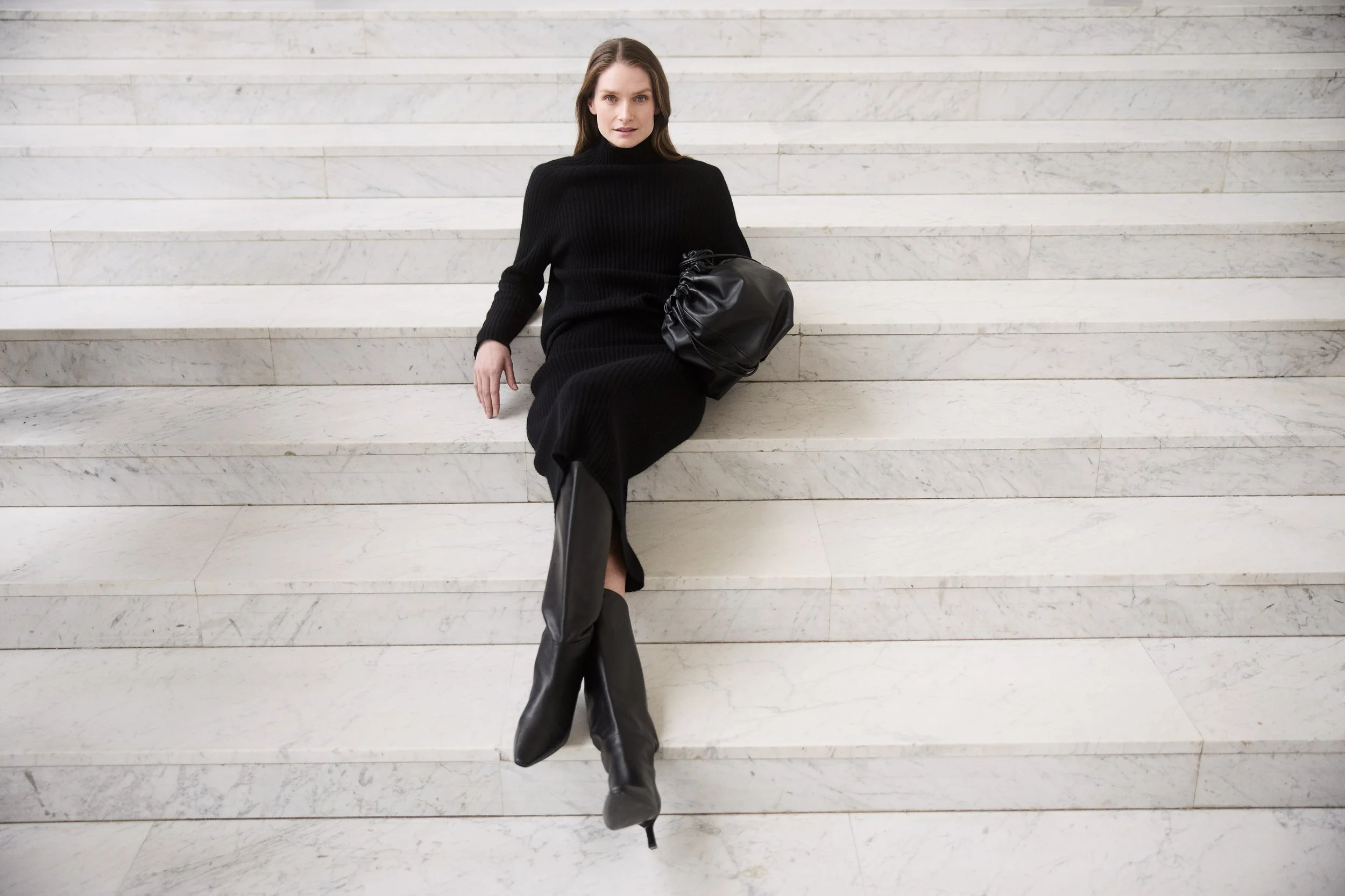 A woman dressed in a black outfit sitting on white marble stairs, holding a black handbag and wearing black high-heeled boots.