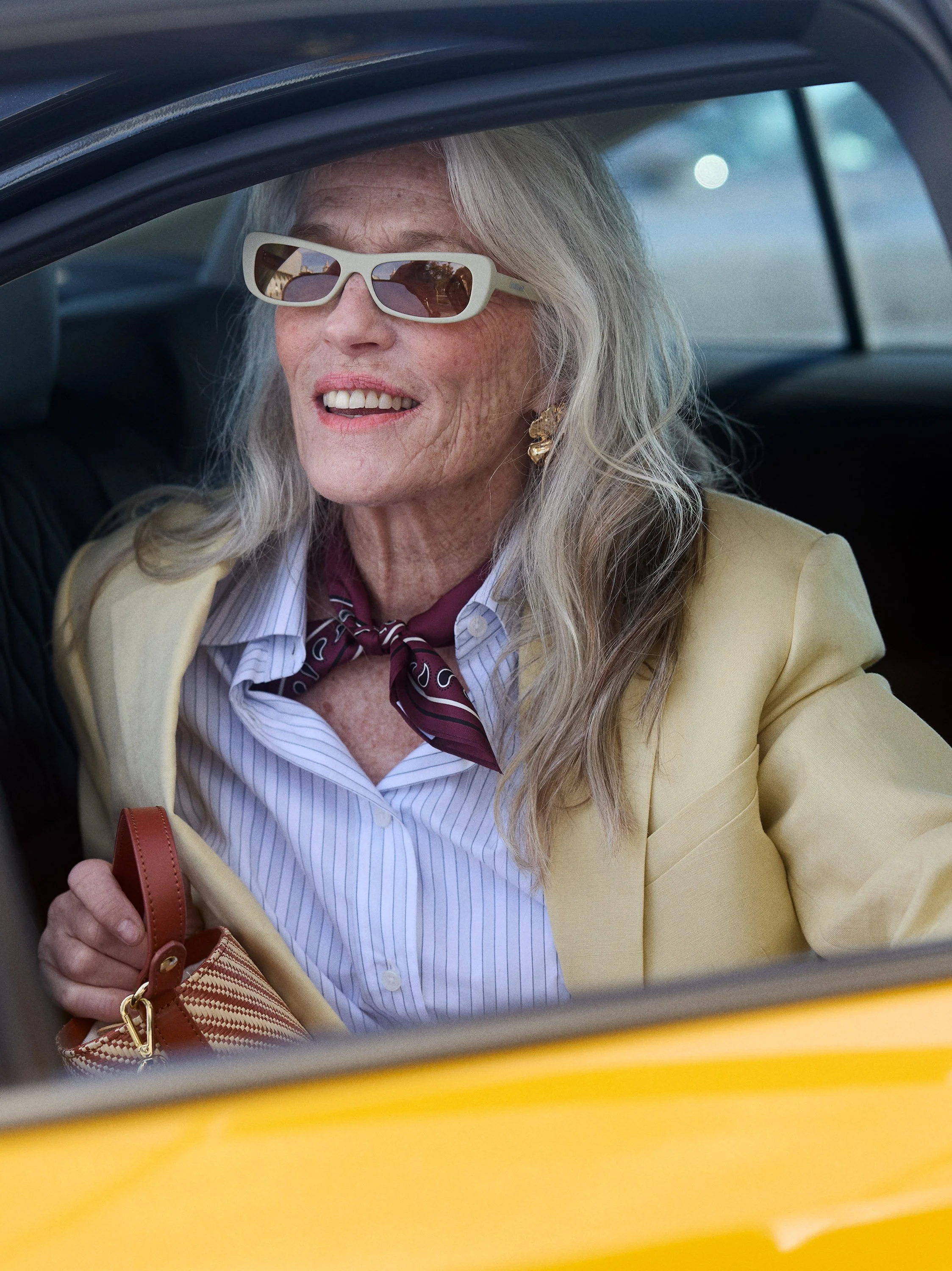 An elderly woman with long gray hair wearing sunglasses, a yellow blazer, a striped shirt, a maroon neck scarf, and gold earrings, smiling as she sits in the back seat of a yellow car.