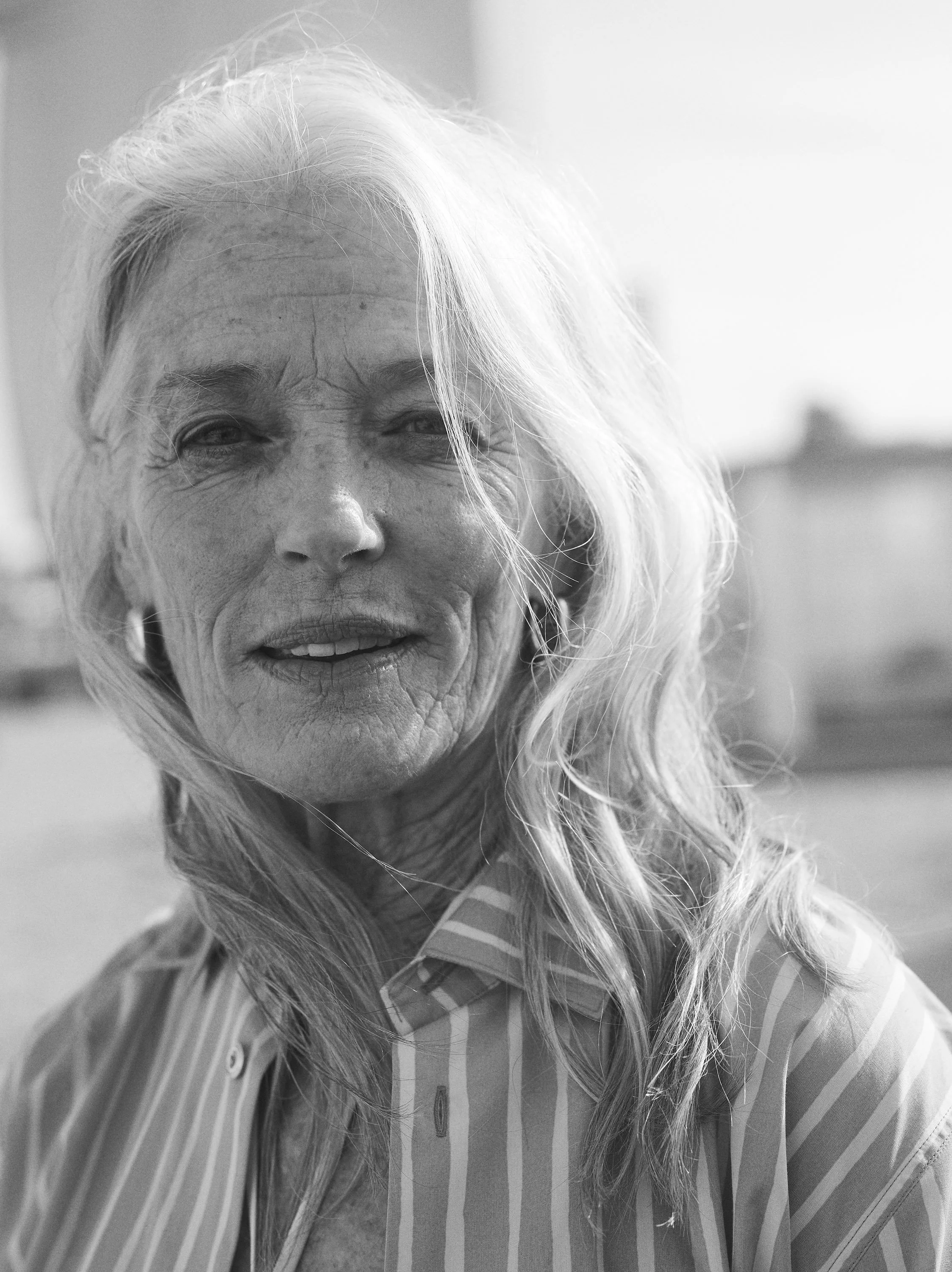 Close-up black and white photo of an elderly woman with long wavy hair, wearing a striped shirt, smiling outdoors.