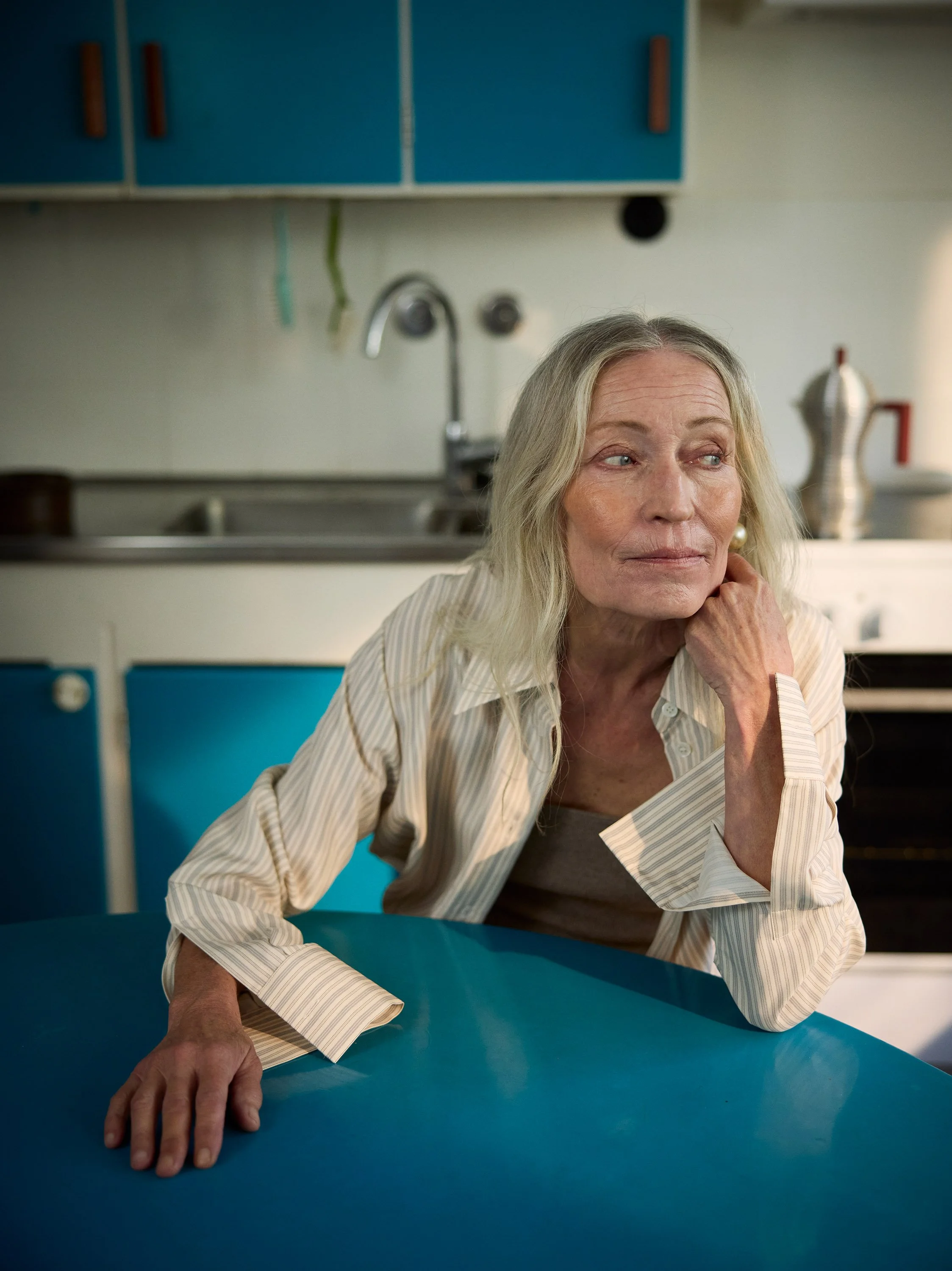 An elderly woman with long gray hair sitting at a blue kitchen table, looking thoughtfully to the side. The kitchen has white walls, blue cabinets, a stainless steel sink, and some kitchen utensils.