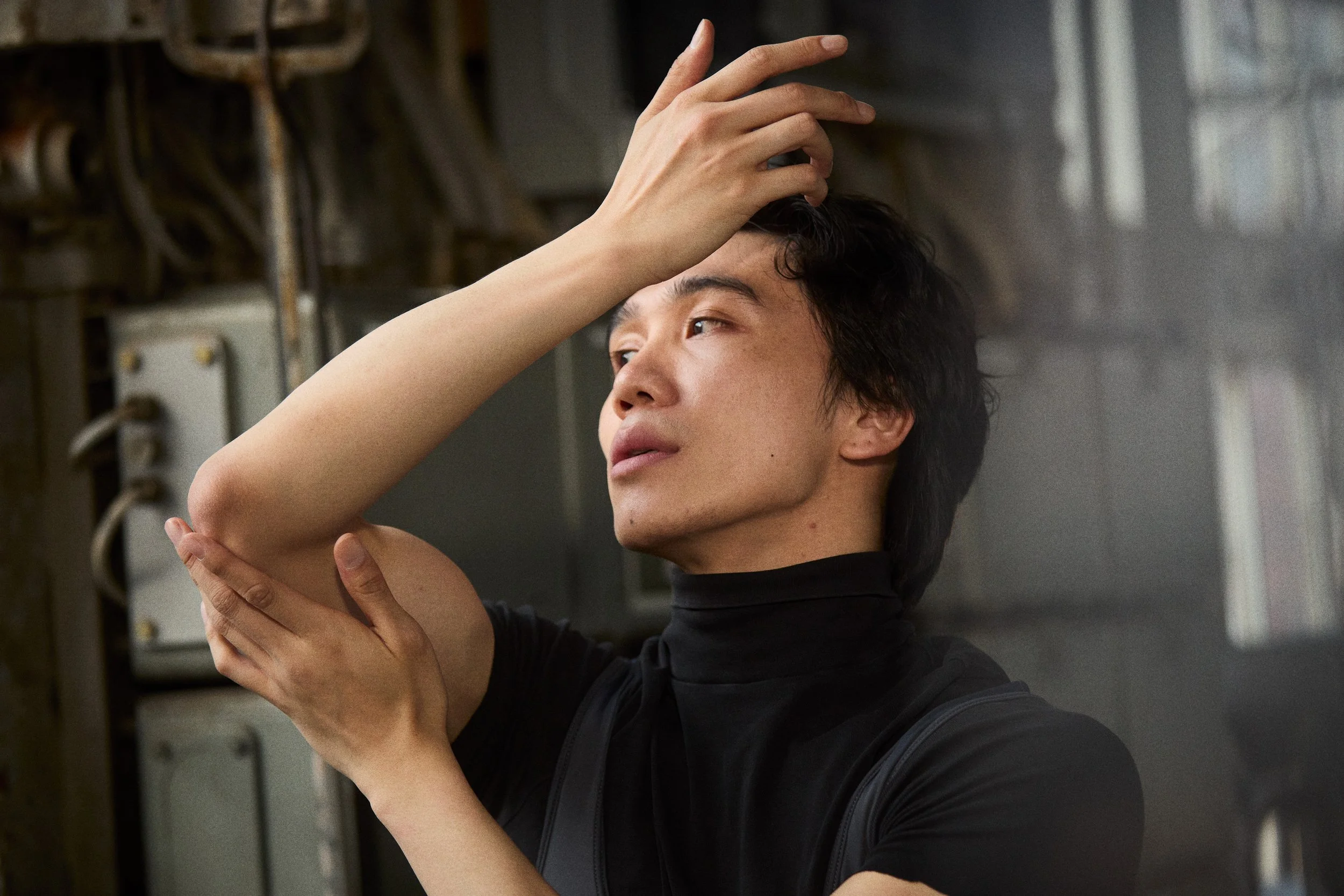 A person with short dark hair wearing a black turtleneck poses with one hand on their forehead and the other on their shoulder, in an industrial setting.
