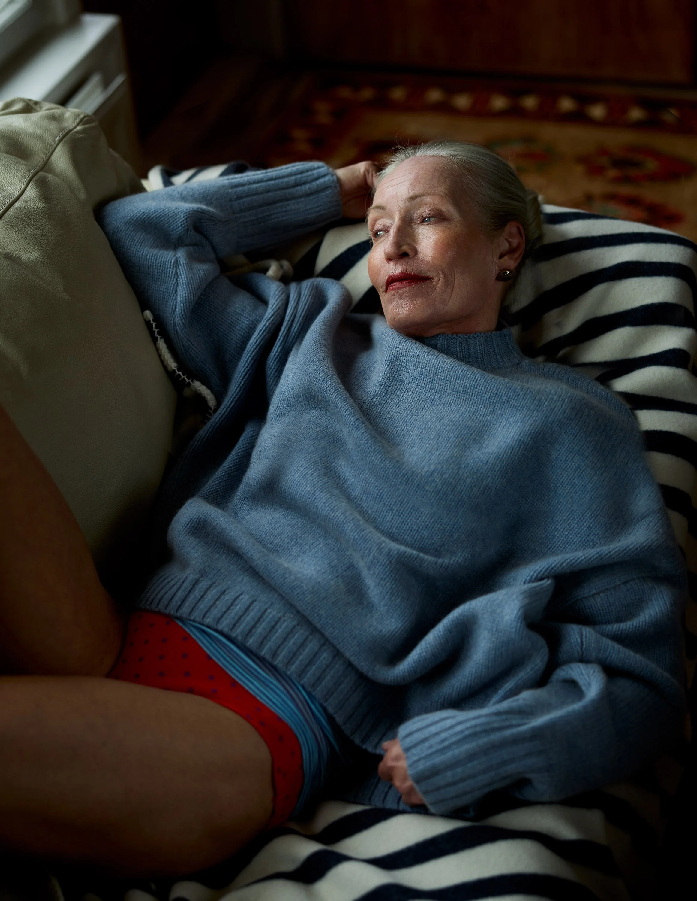 An older woman relaxing on a striped pillow, wearing a blue sweater and red polka dot underwear, lying on her side on a couch or bed with a patterned rug in the background.