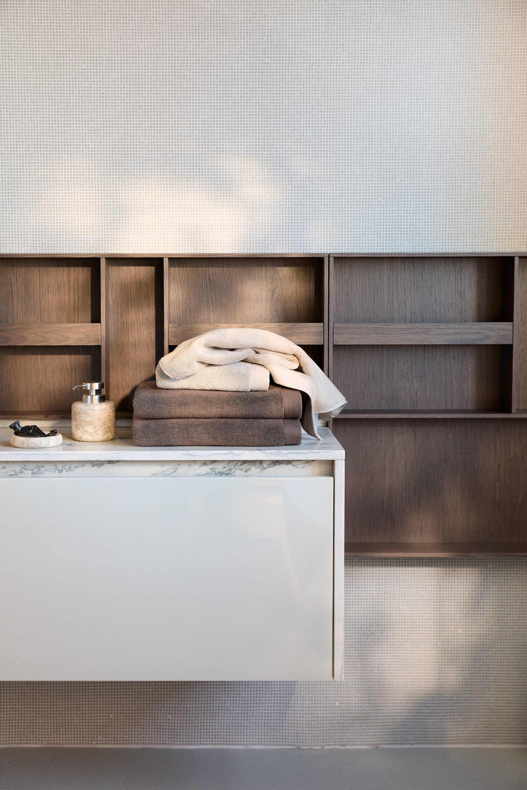 A modern bathroom cabinet with folded towels and toiletries on a marble countertop, wooden shelves in the background, and soft lighting.