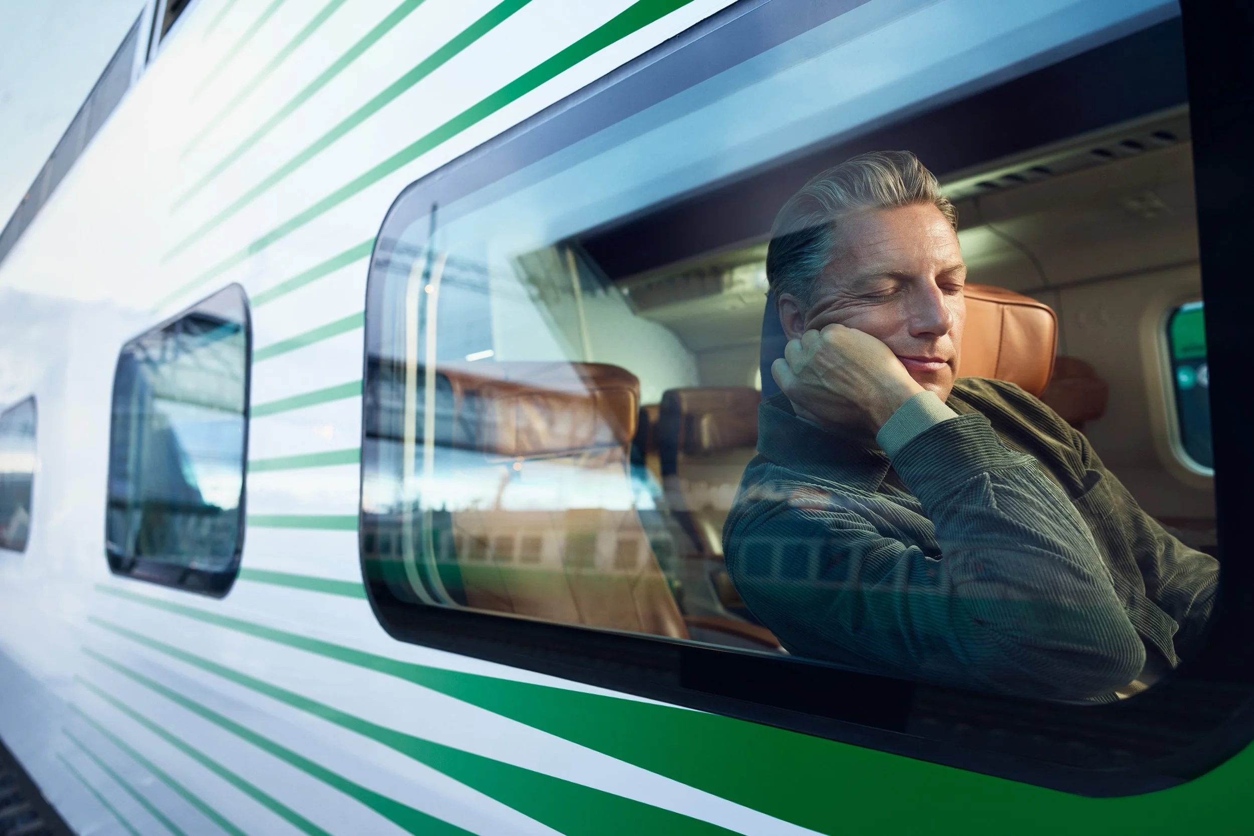 A man sitting on a train, resting with his eyes closed, appears to be sleeping or relaxing inside a train car with brown seats, as seen through a window with reflections.
