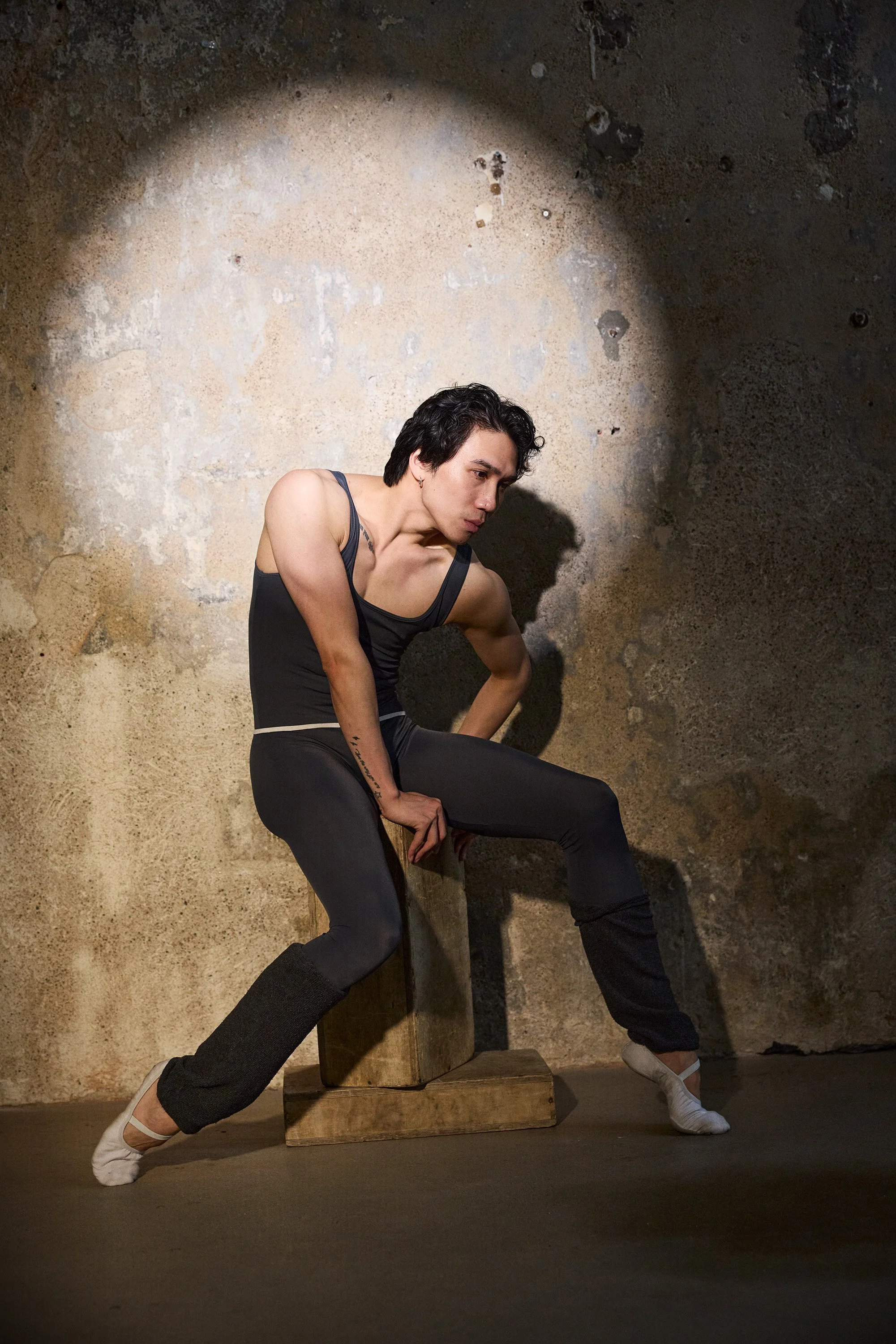 Male dancer in black dancewear posing outdoors against a weathered wall, sitting on a wooden box, with dramatic lighting.