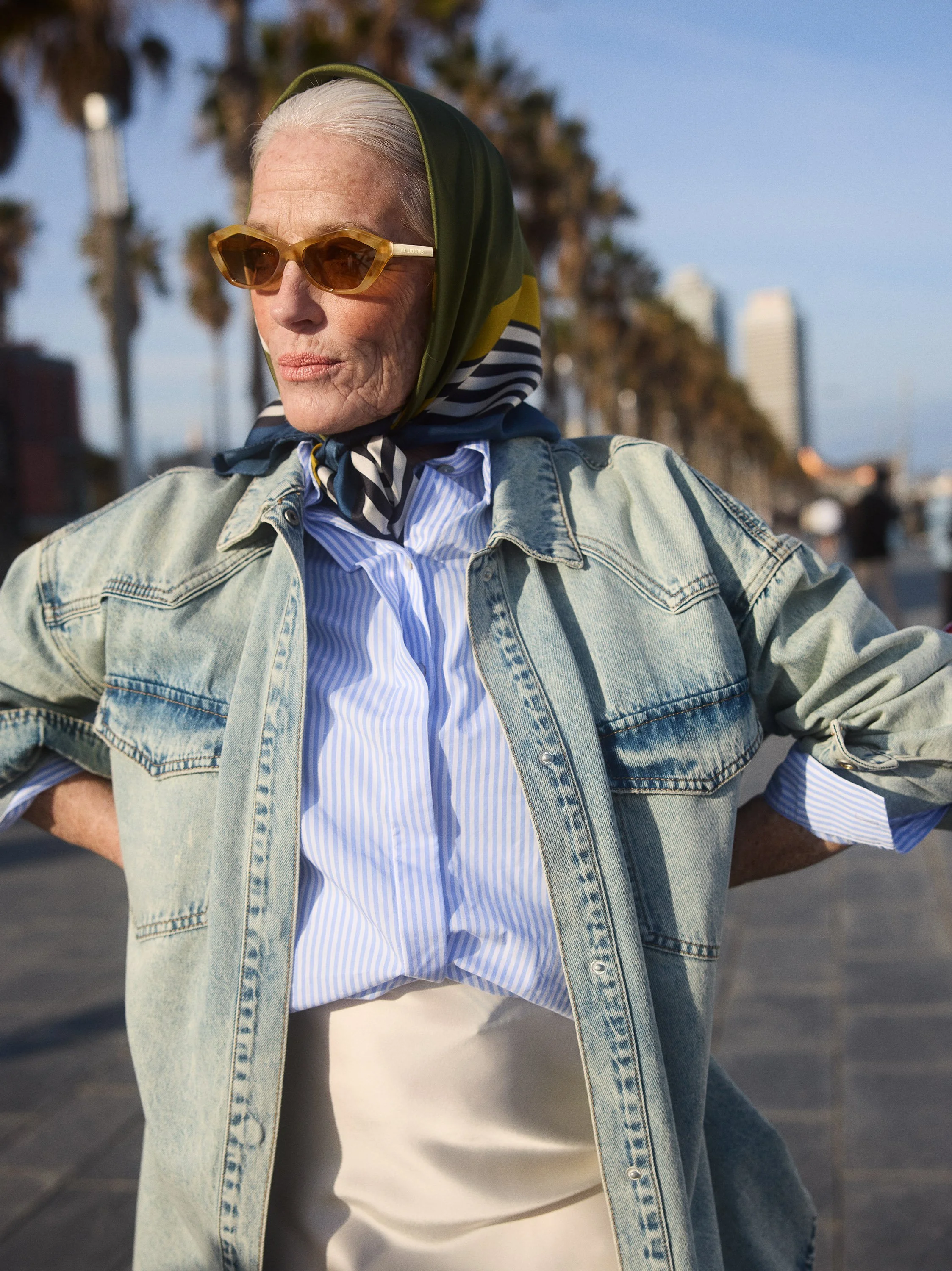 Older woman wearing sunglasses, a patterned headscarf, a striped shirt, a denim jacket, and a cream skirt stands outdoors on a sunny day with palm trees and city buildings in the background.