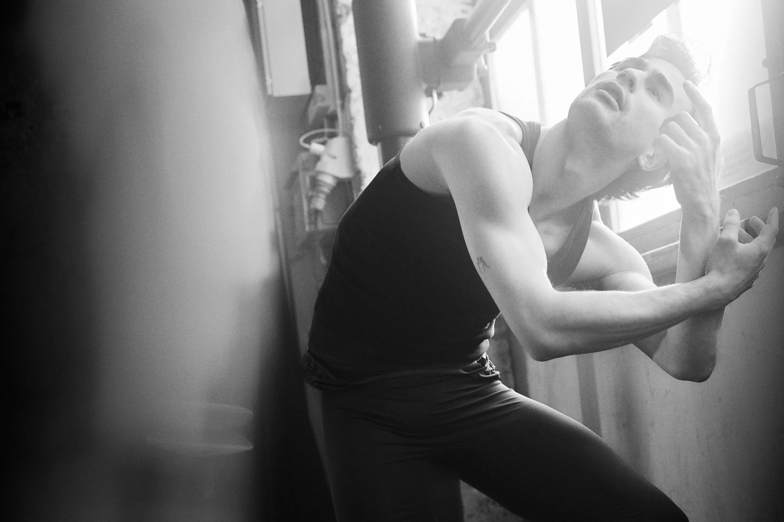 A person in a sleeveless top leaning against a wall near a window, with hand on forehead, appearing to be in a reflective or emotional state.