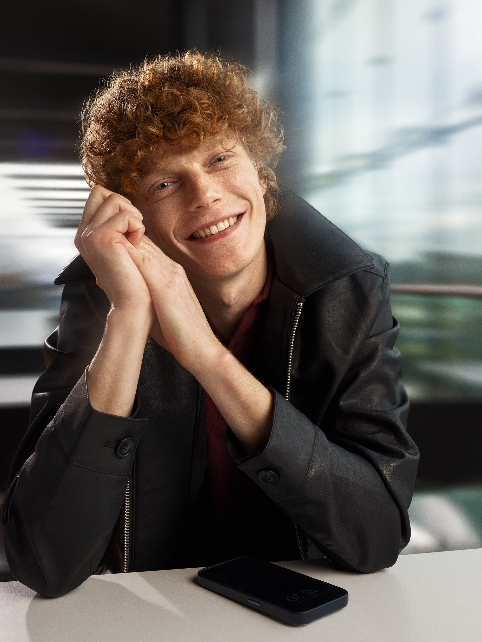 A young man with curly red hair and a broad smile, wearing a black leather jacket, sitting at a table with a smartphone in front of him, in a modern indoor setting.