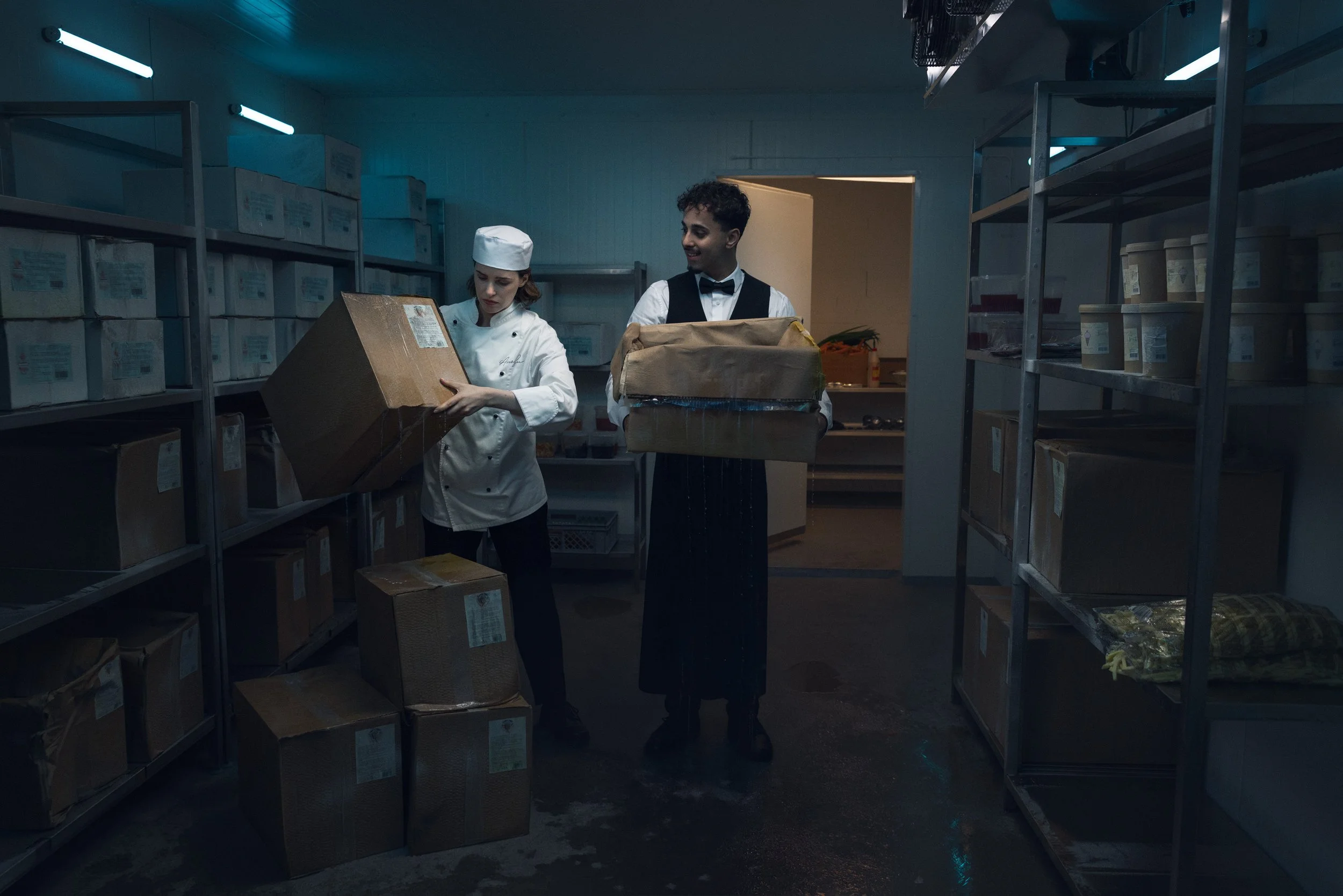 A chef and a waiter standing in a storage room with shelves full of boxes, with the chef handling a box and the waiter holding another box.