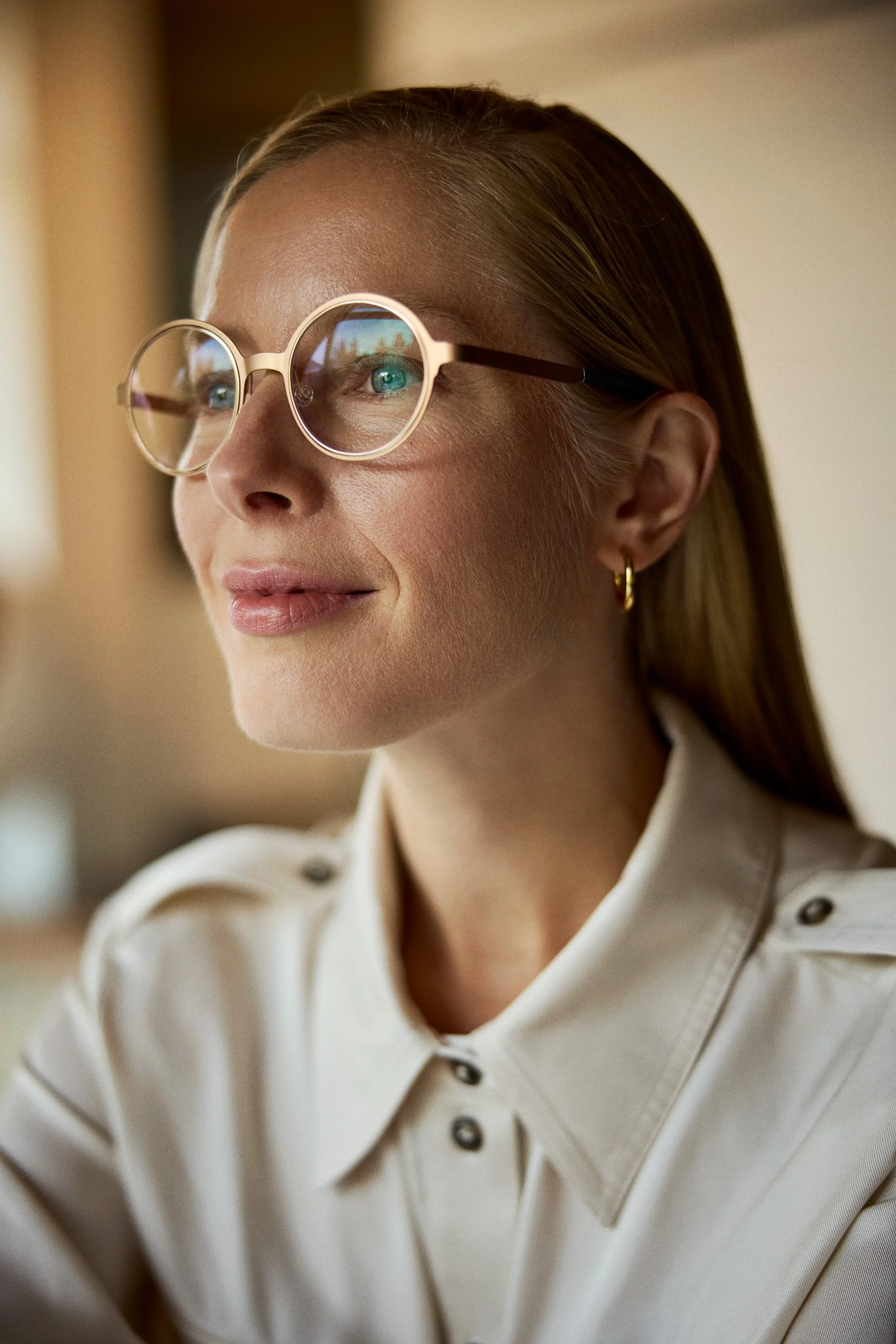 Close-up of a woman wearing large round glasses, gold hoop earrings, and a beige shirt with button details, smiling softly.