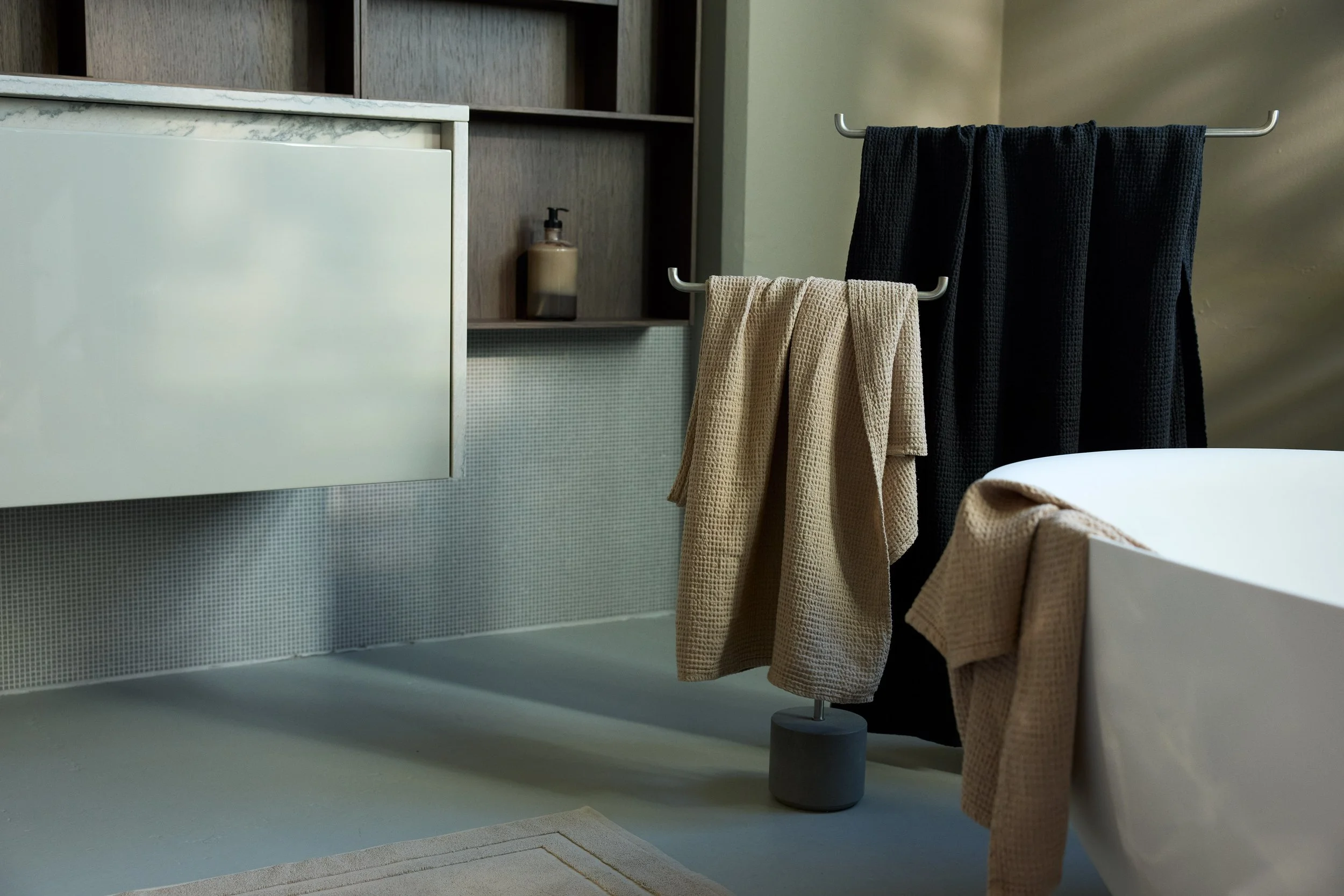 Modern bathroom with beige and black towels hanging on a silver towel rack, a white bathtub, and a small white cabinet with a soap dispenser on a wooden shelf in the background.