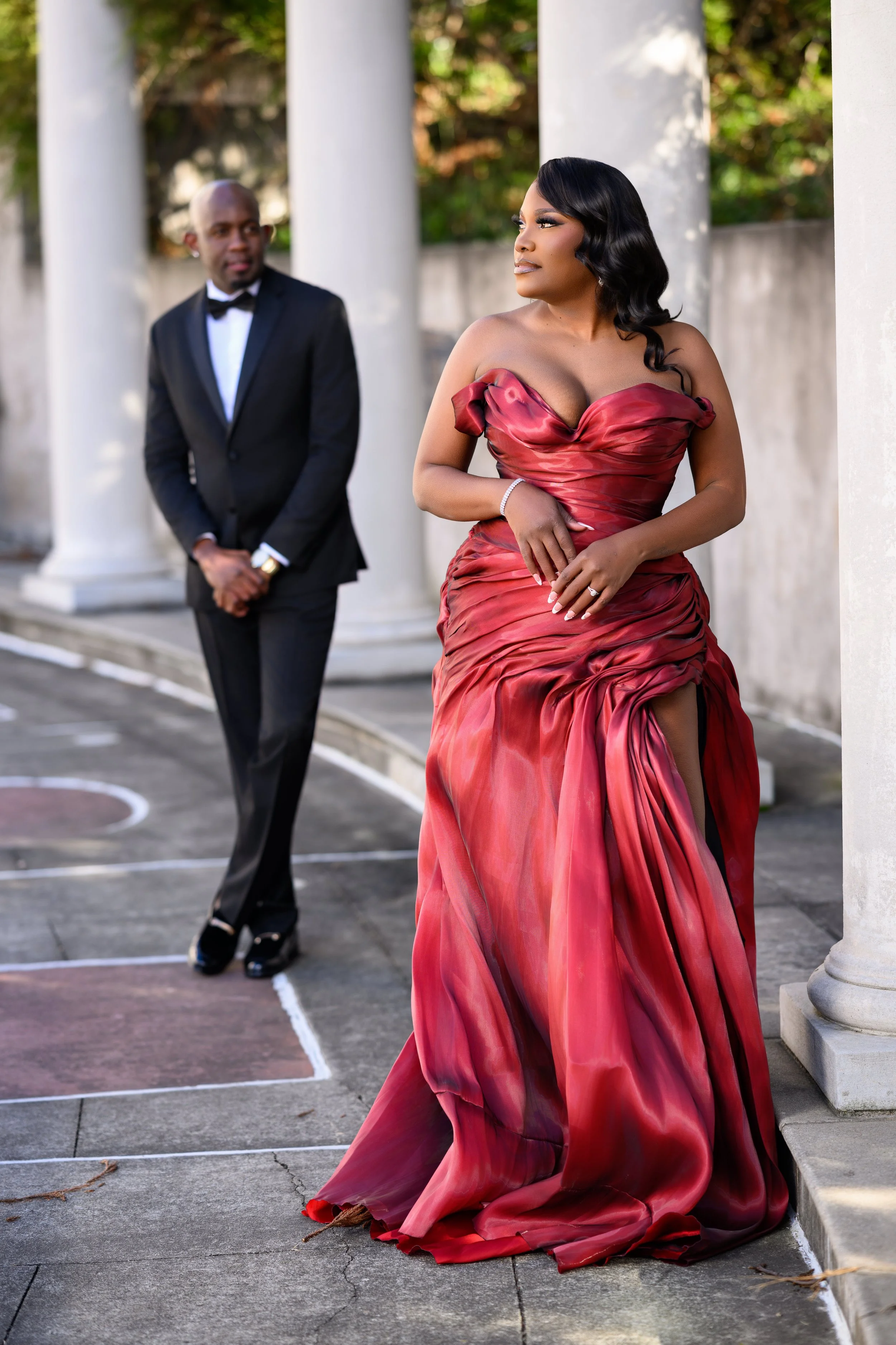 Fashion-forward engagement photo of a woman in a dramatic red gown walking ahead while her partner looks on at Millennium Gate Museum.