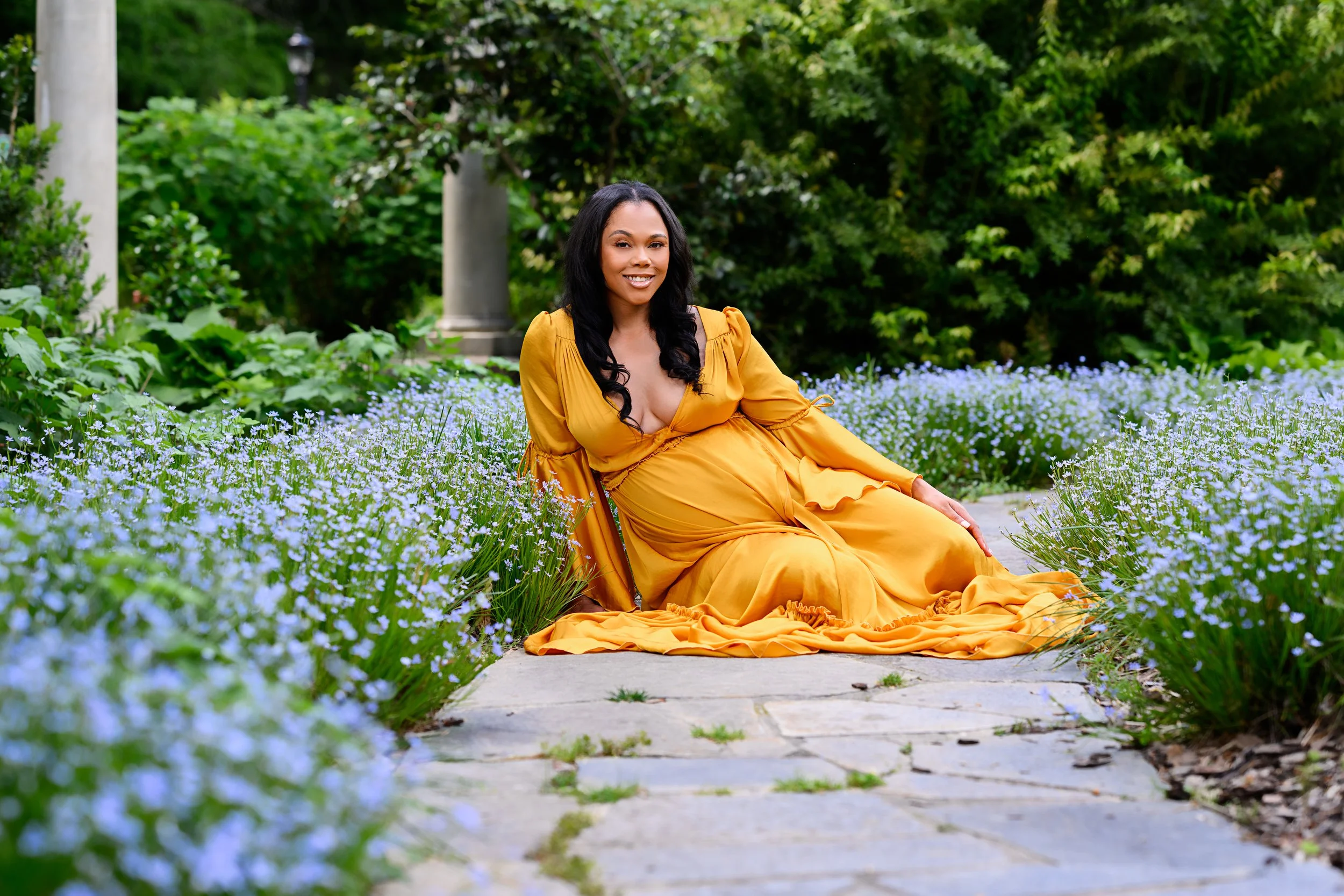 Expectant mother seated among blue flowers in a flowing golden gown during her Atlanta maternity session.
