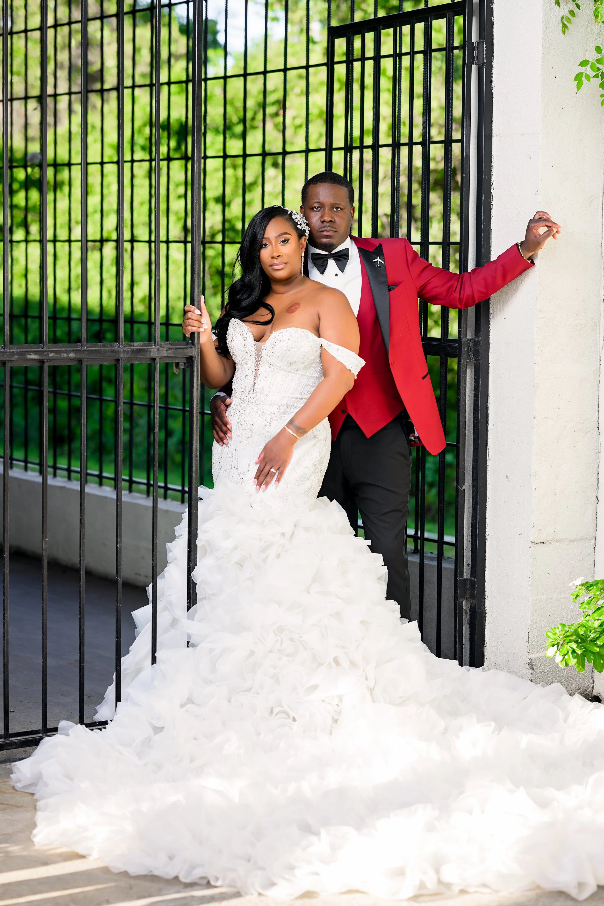 Editorial-style wedding portrait of a Black couple in formal attire standing at a garden gate, featuring a flowing bridal gown, modern styling, and natural greenery.