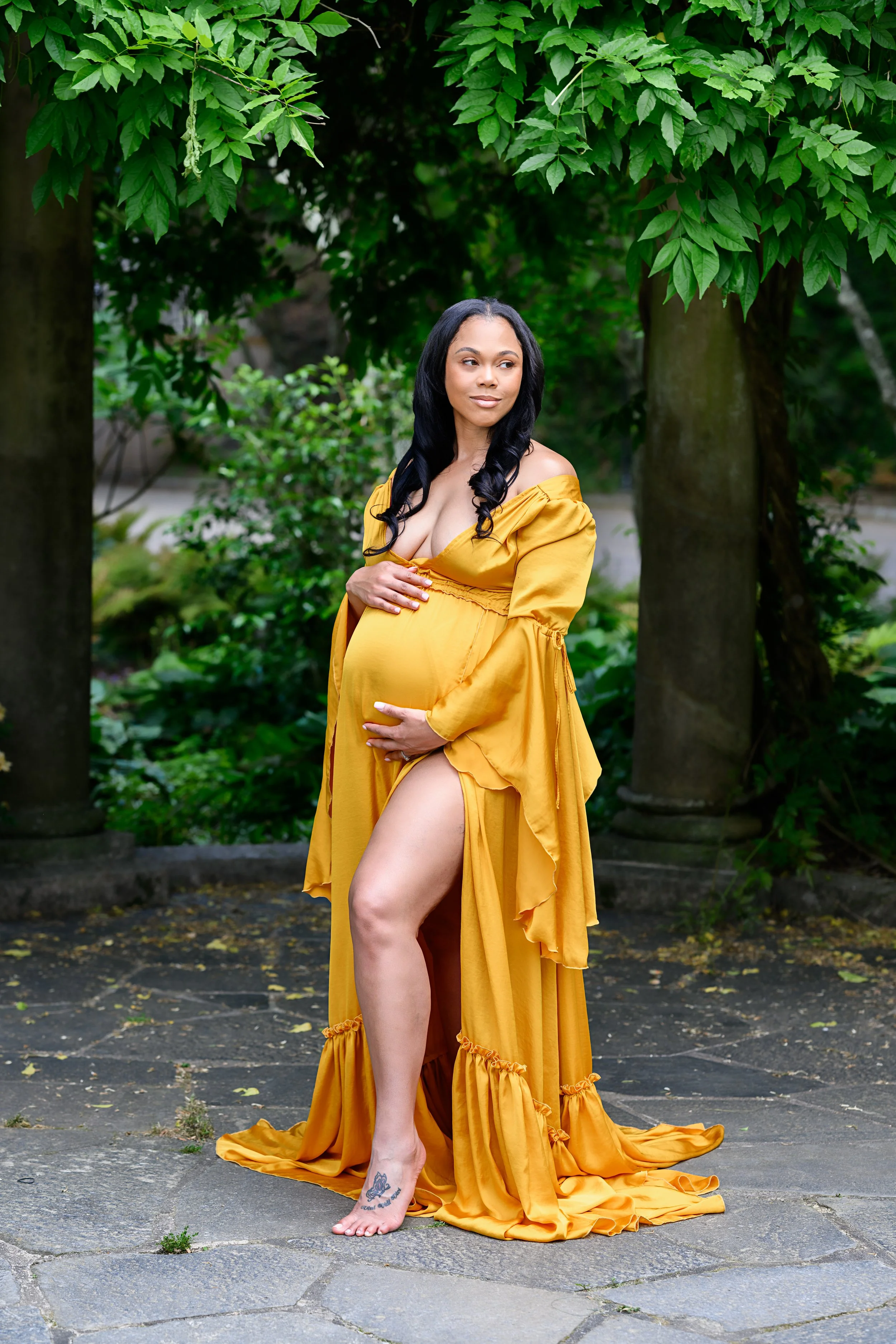 Mother-to-be posing in a golden maternity gown beneath lush green trees in Atlanta.