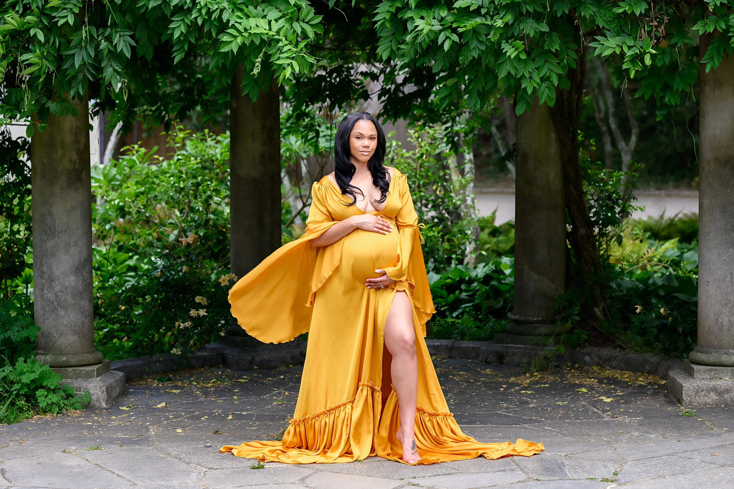 Expectant mother in a flowing golden maternity gown surrounded by lush greenery during an outdoor maternity session in Atlanta.