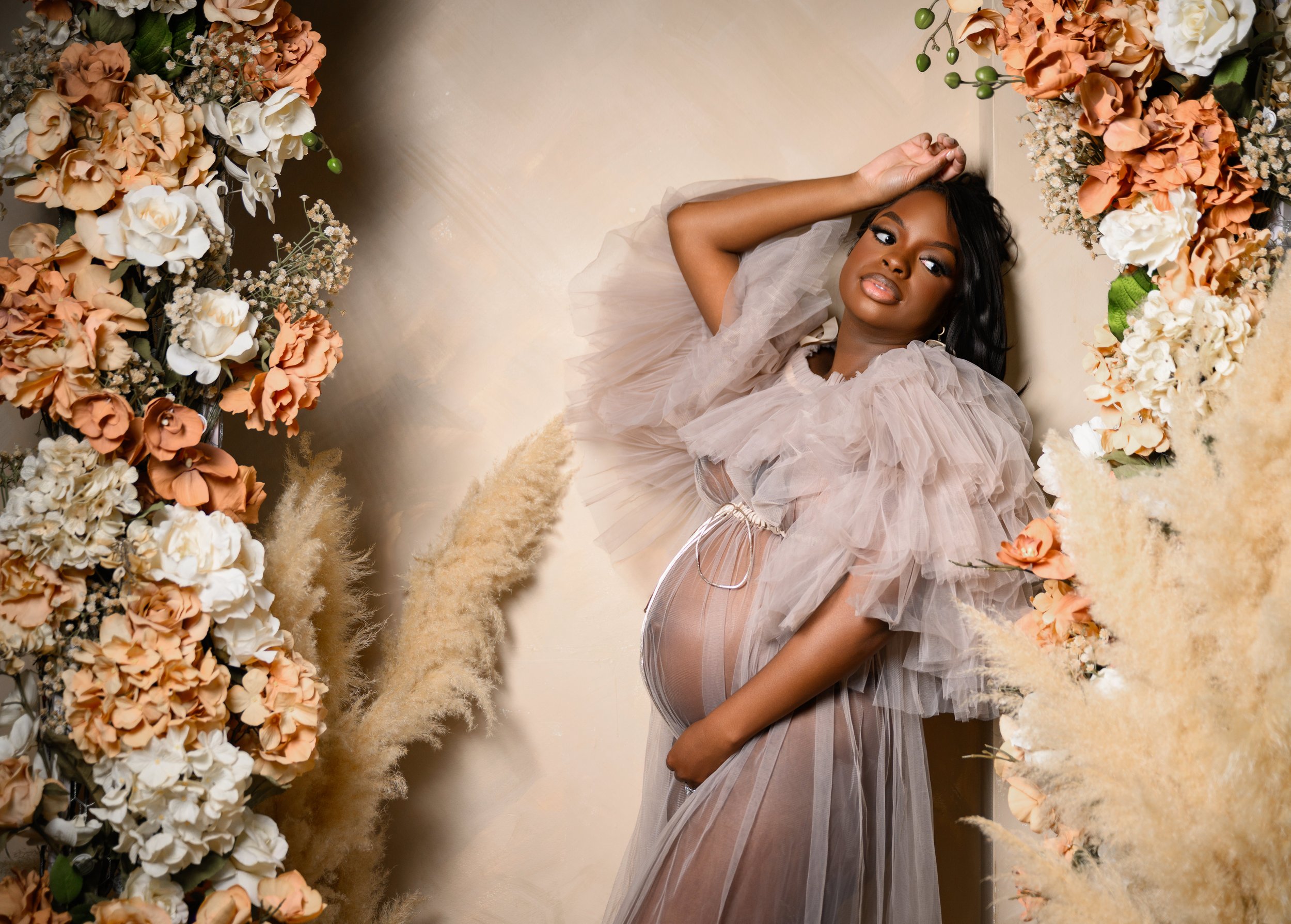 Expecting mother posing in a soft neutral maternity gown surrounded by warm floral details in an Atlanta studio.