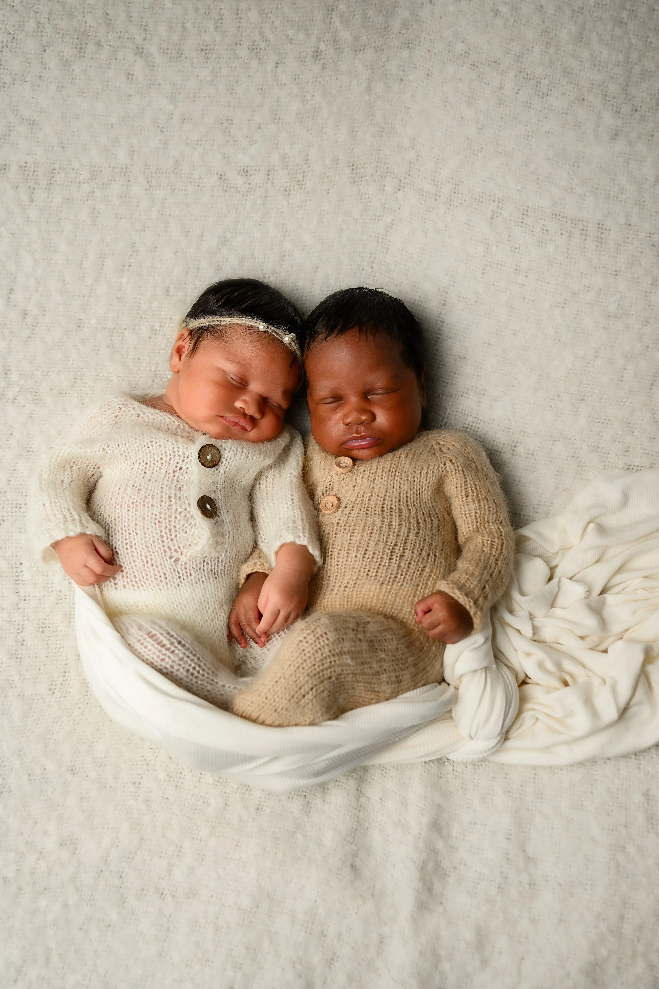 Overhead portrait of newborn twins sleeping together in coordinating neutral knit outfits.