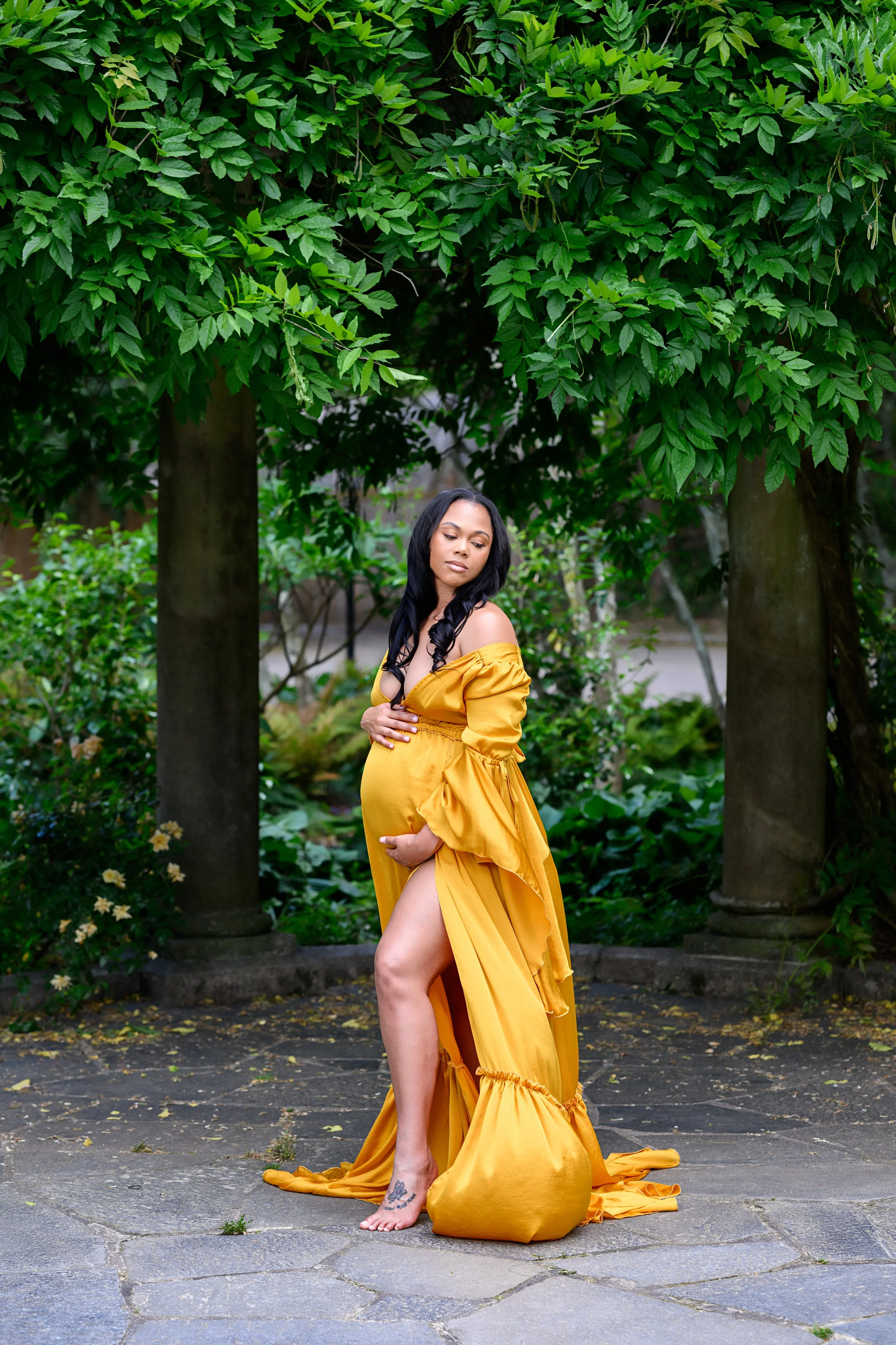 Side-profile maternity portrait of an expectant mother cradling her baby bump in a golden gown surrounded by greenery.