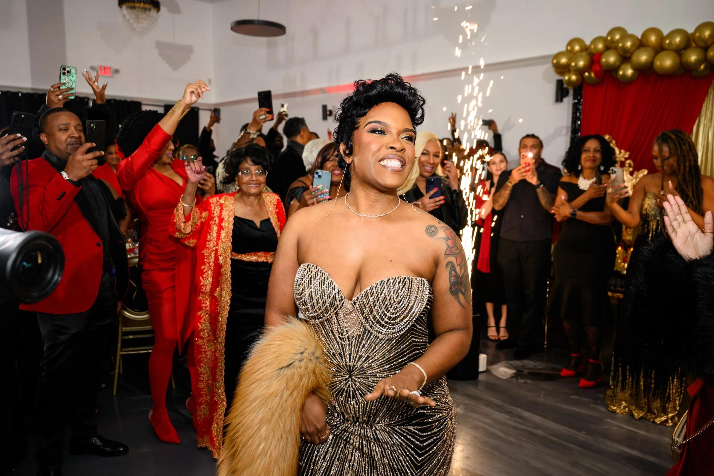 Woman making her entrance during a birthday celebration surrounded by guests and indoor sparkler effects.
