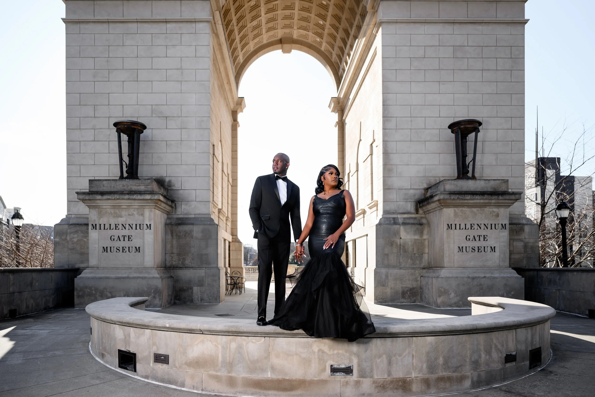 Stylish couple holding hands during an engagement session beneath the Millennium Gate Museum arch in Atlanta on a bright winter afternoon.
