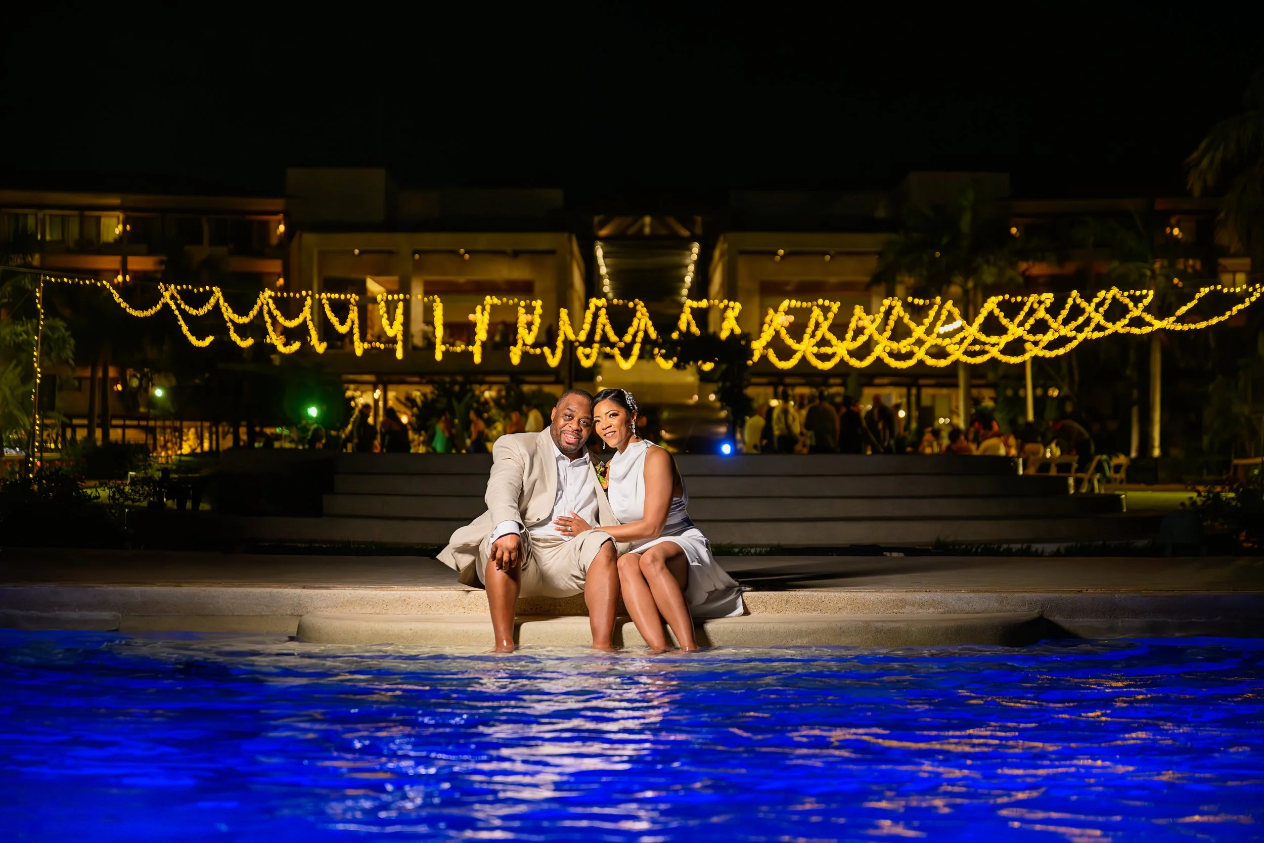 Tekla and Rick sitting poolside under string lights during their destination wedding celebration in Cancun, Mexico.