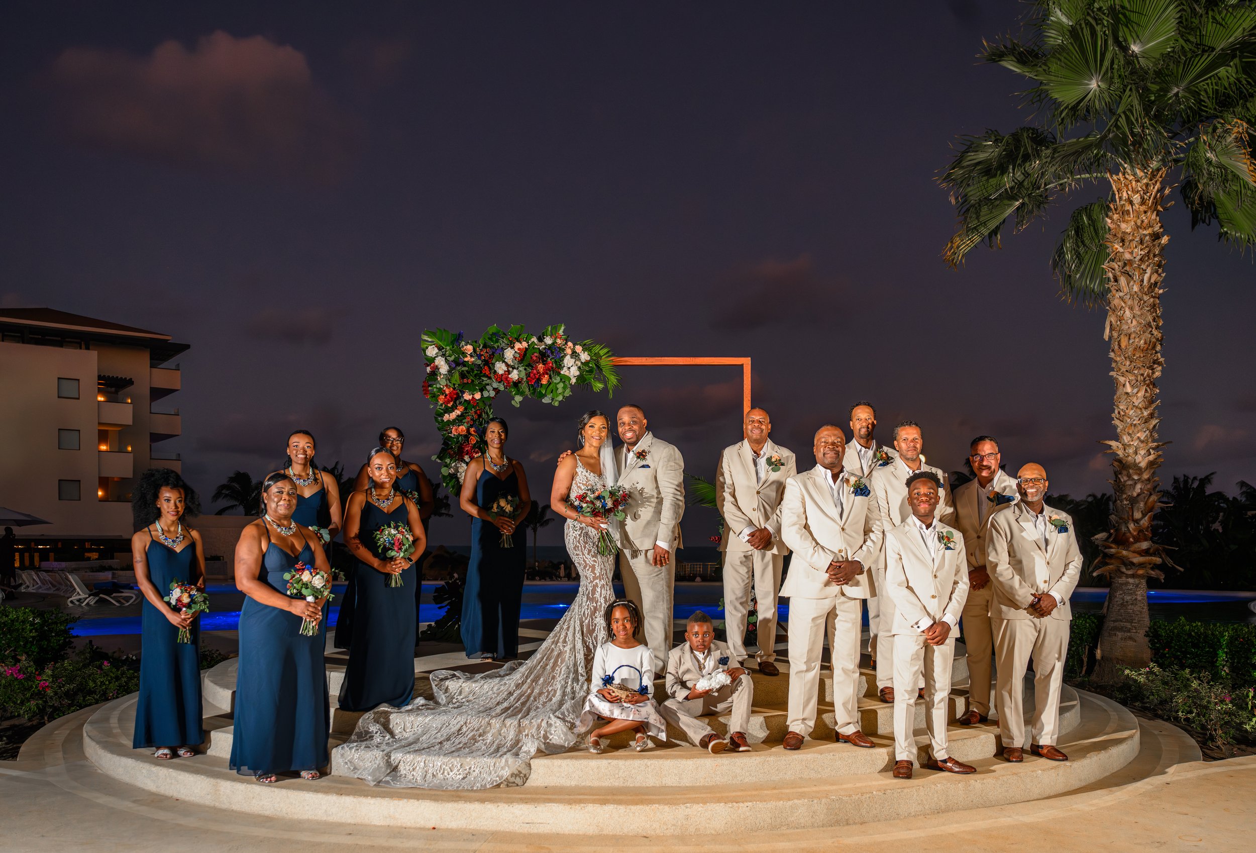 Tekla and Rick with their wedding party beneath the floral ceremony arch at dusk in Cancun.