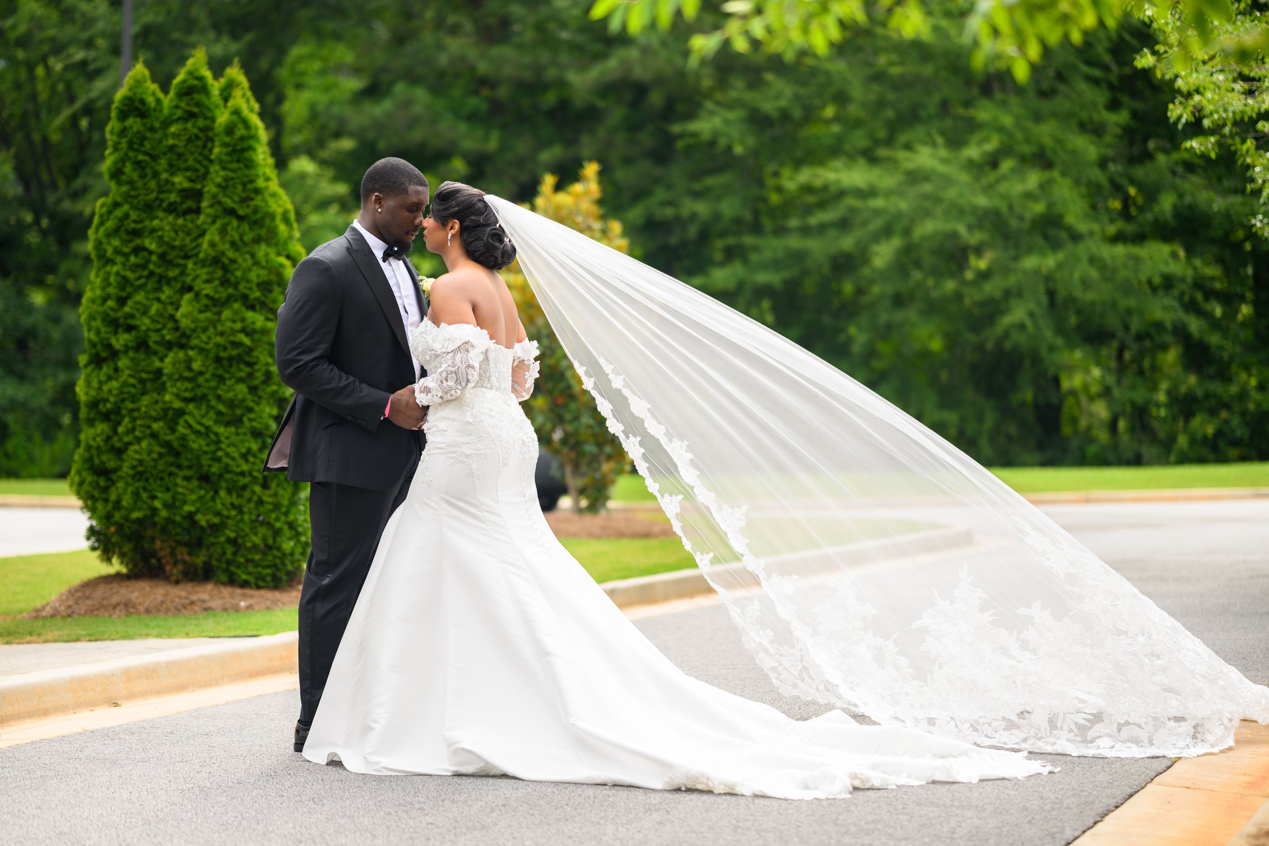 Bride and groom posing outdoors with the bride’s long lace veil flowing behind them at The Venue at Daisy Hill.