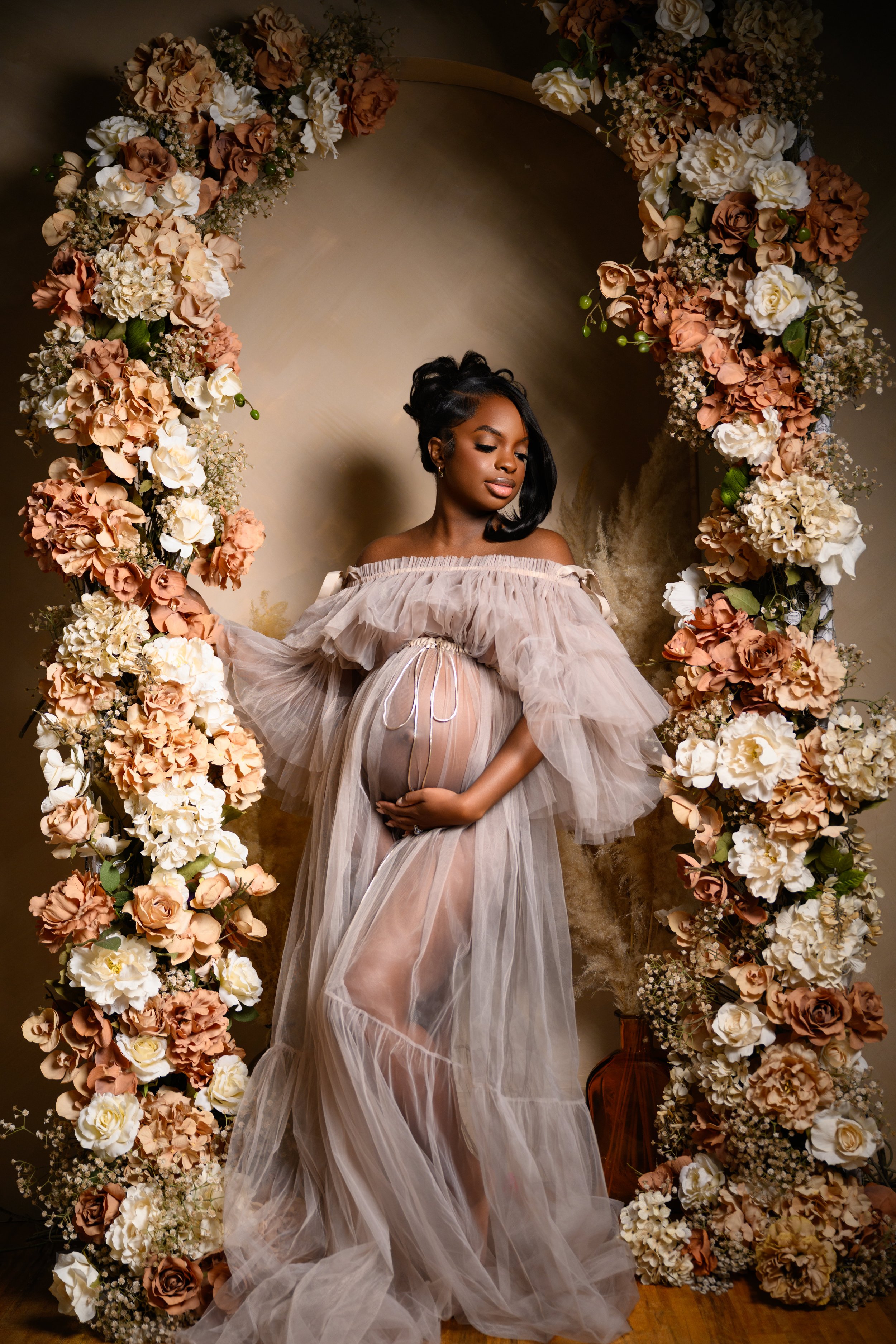 Expecting mother posing in a sheer neutral maternity gown surrounded by a floral arch during a Midtown Atlanta studio maternity session.