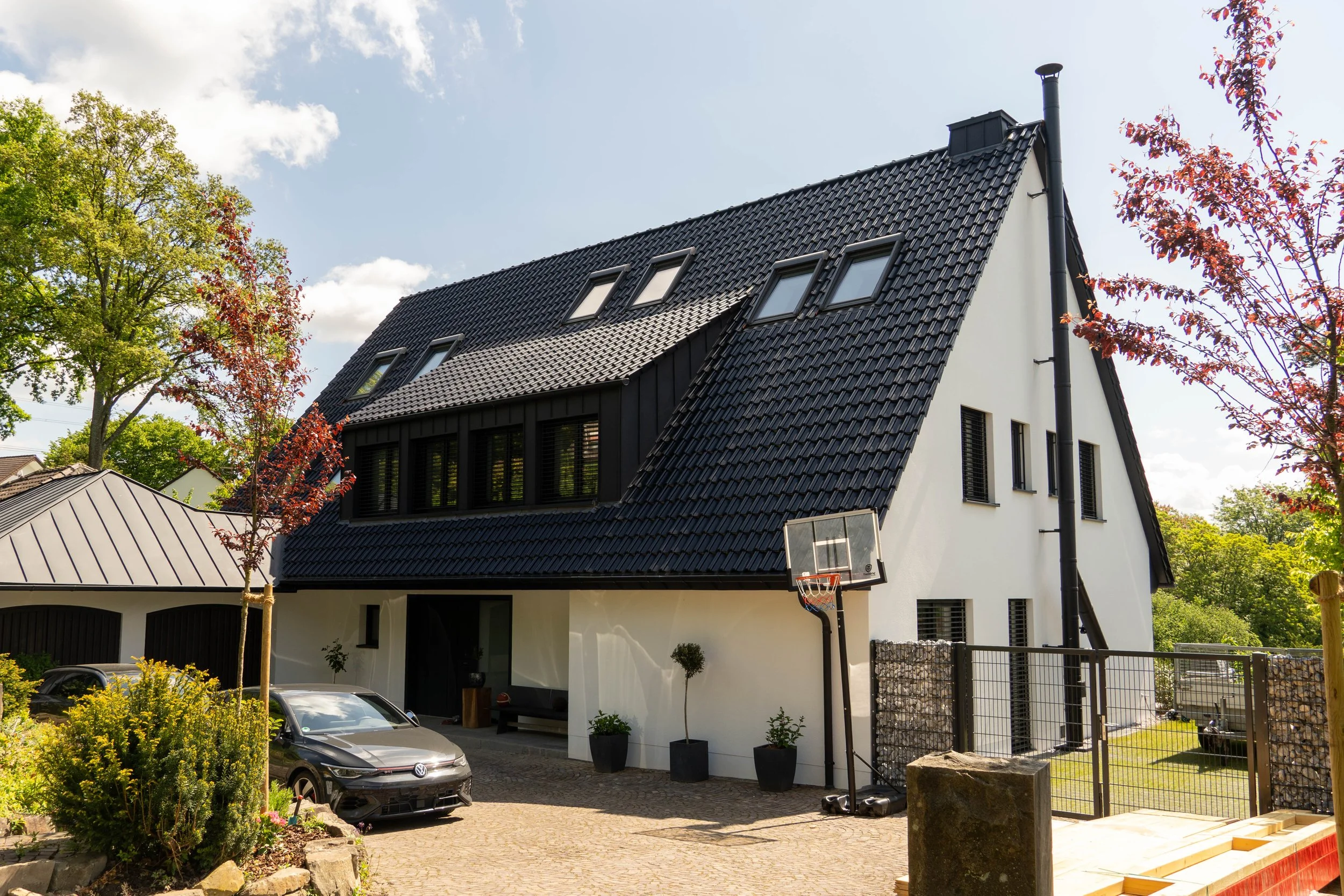 Modernes zweistöckiges Haus mit dunklem Satteldach, Dachfenstern und weißen Wänden, umgeben von Bäumen und einem Garten, mit Parkplätzen und einem Basketballkorb im Vordergrund.