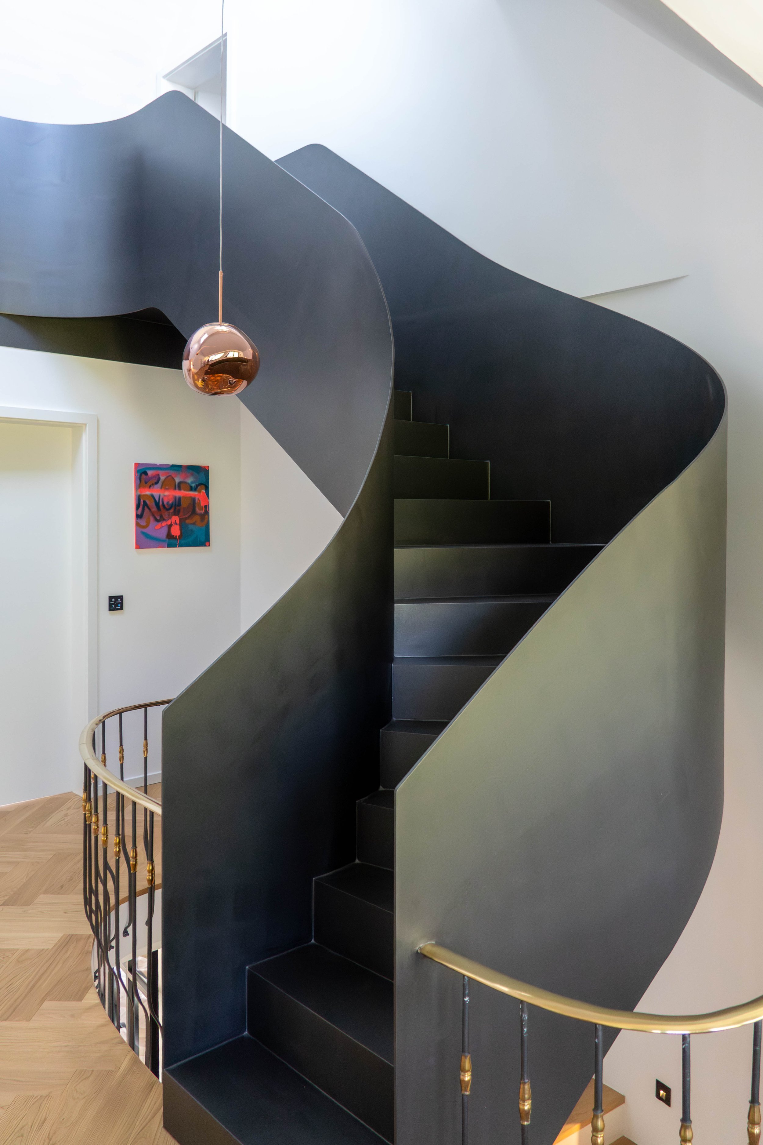 Moderne spiralförmige Treppe mit schwarzer Oberfläche, goldenen Geländern und einer hängenden Kupferlampe in einem hellen, eleganten Raum. Eine bunte abstrakte Malerei hängt an der Wand.