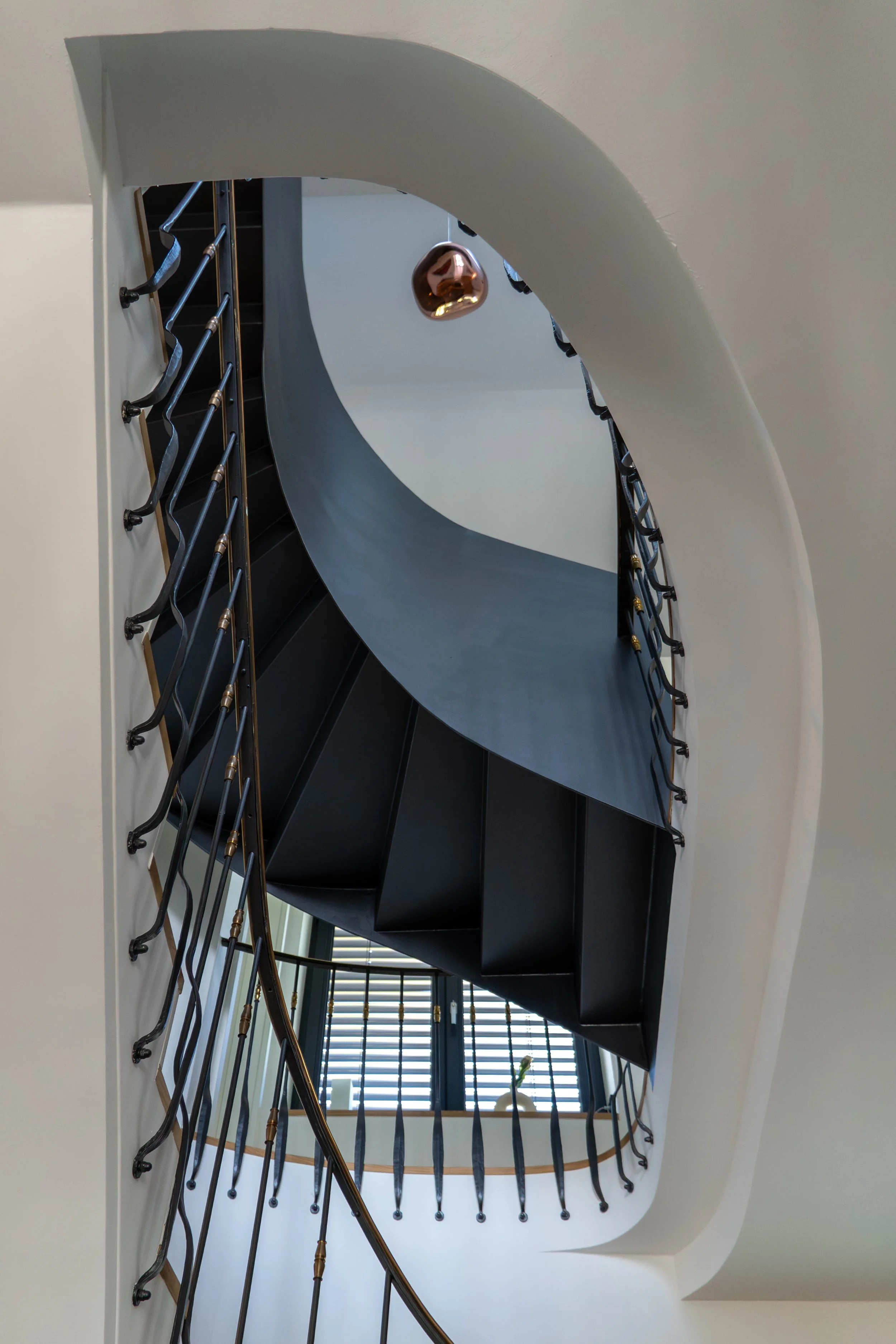 Blick nach oben auf eine spiralförmige schwarze Treppe mit einem gekrümmten Geländer aus Metall und Holzhandlauf, vor einem Fenster mit horizontalen Jalousien und einer Pflanze im Hintergrund.