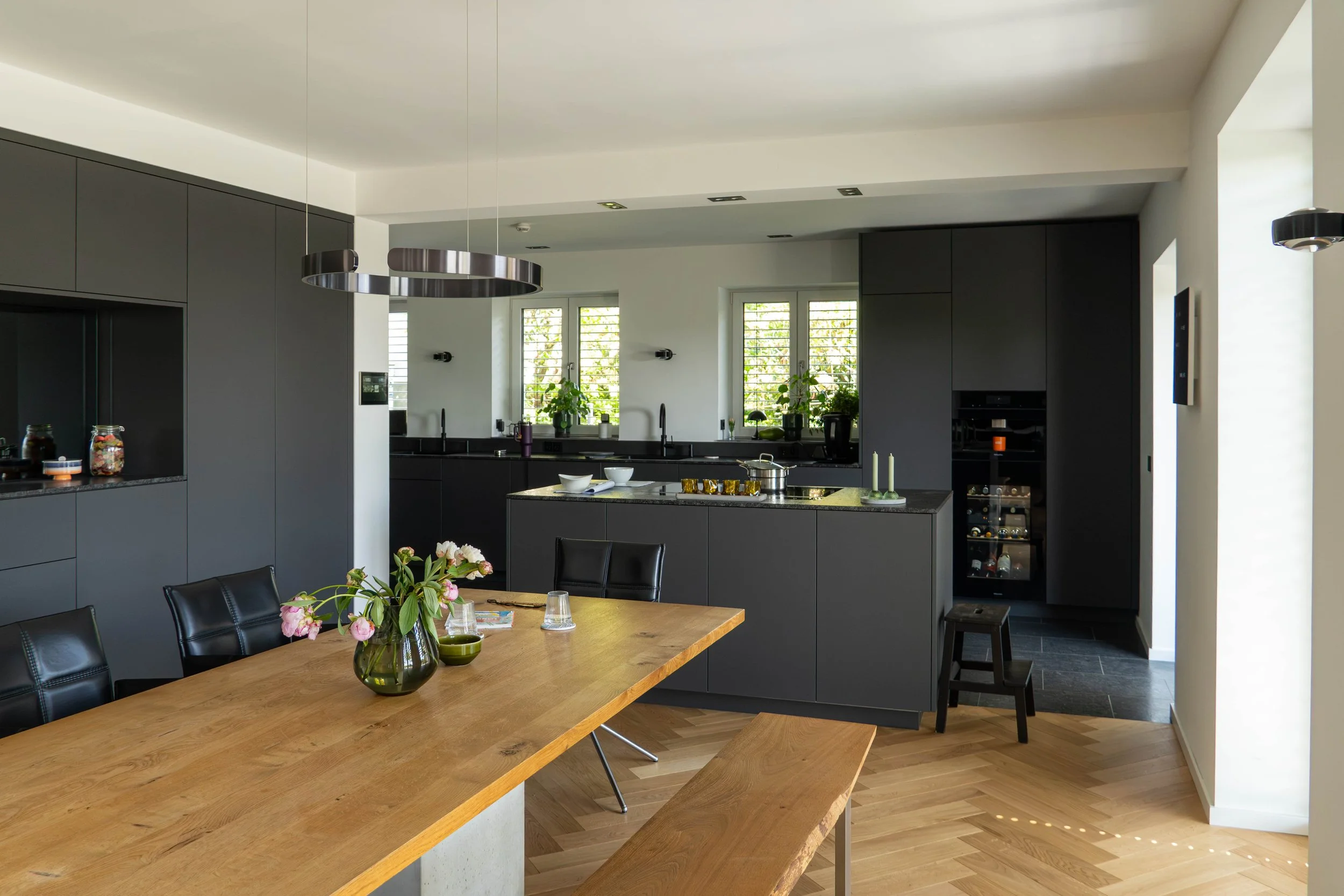 Moderne schwarze Küche mit zentraler Insel, große Fenster mit Lärchenholz-Lamellen, Essbereich mit Holztisch, Blumen im Vase, schwarze Stühle.