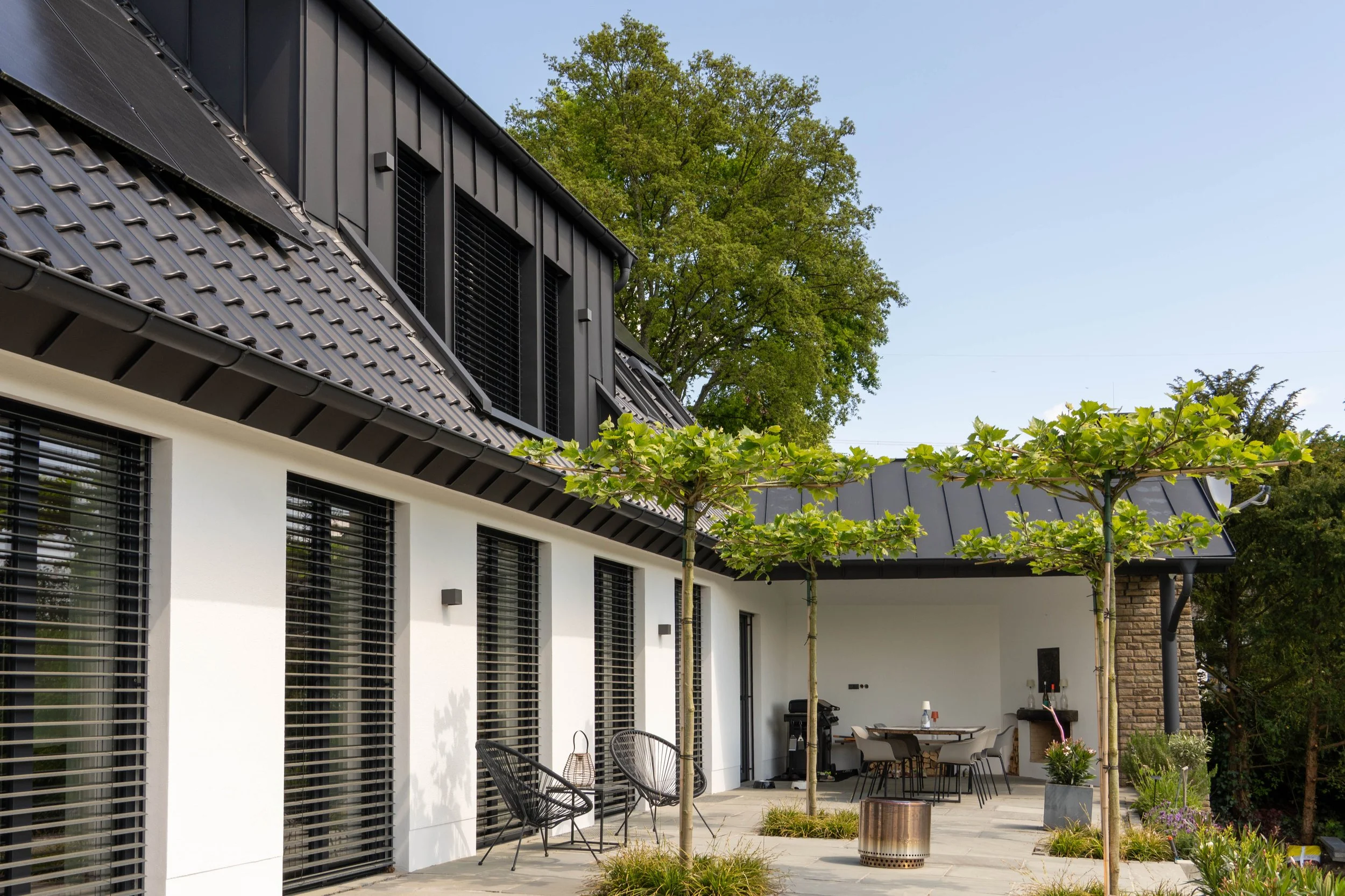 Moderne Hausfassade mit weißen Wänden, großen Fenstern mit Rollläden, einem Balkon mit schwarzen Geländern, grünem Baum und einer Terrasse mit Gartenmöbeln und Pflanzen.