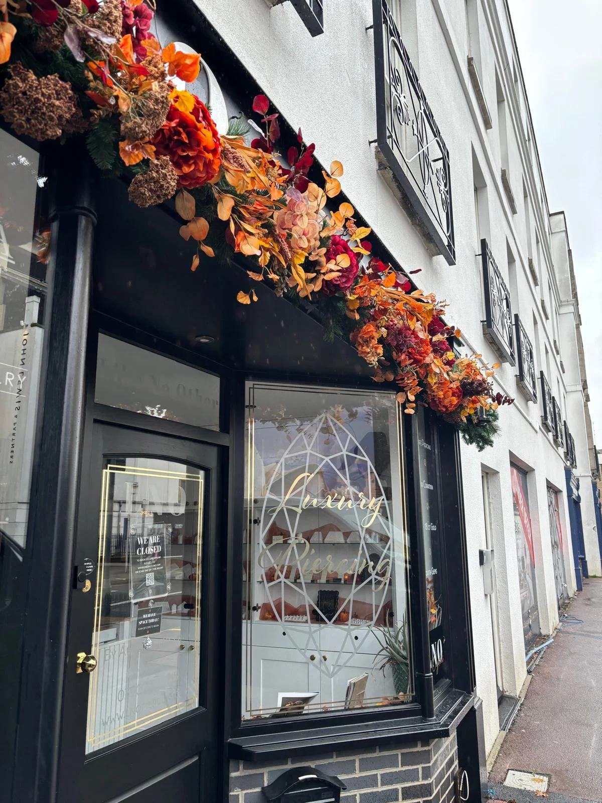 Luxury piercing studio storefront with black trim, geometric window lettering, and an elaborate orange and burgundy floral display.