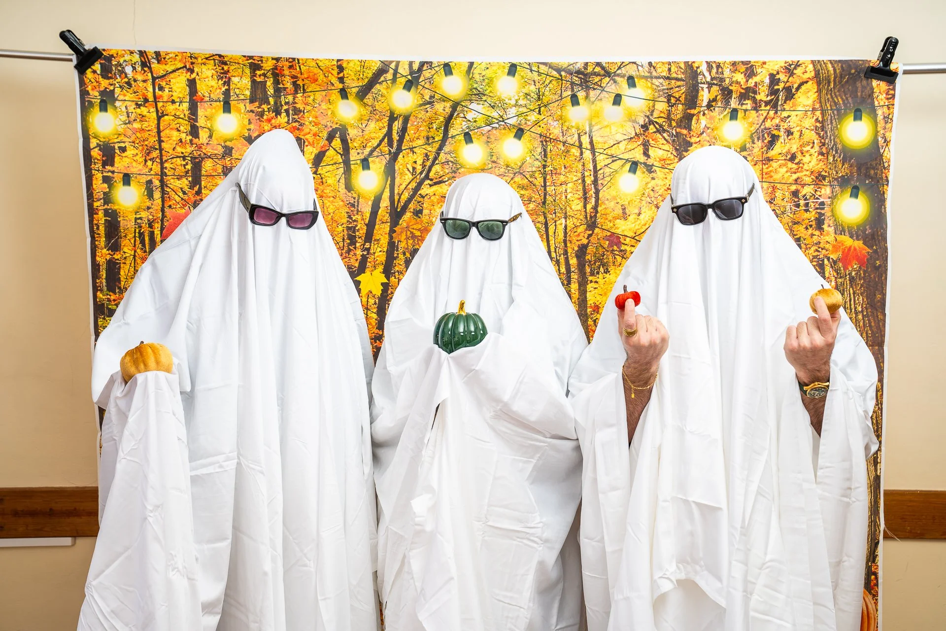 Three people dressed as ghosts in sunglasses pose with pumpkins in front of an autumn forest backdrop and string lights.