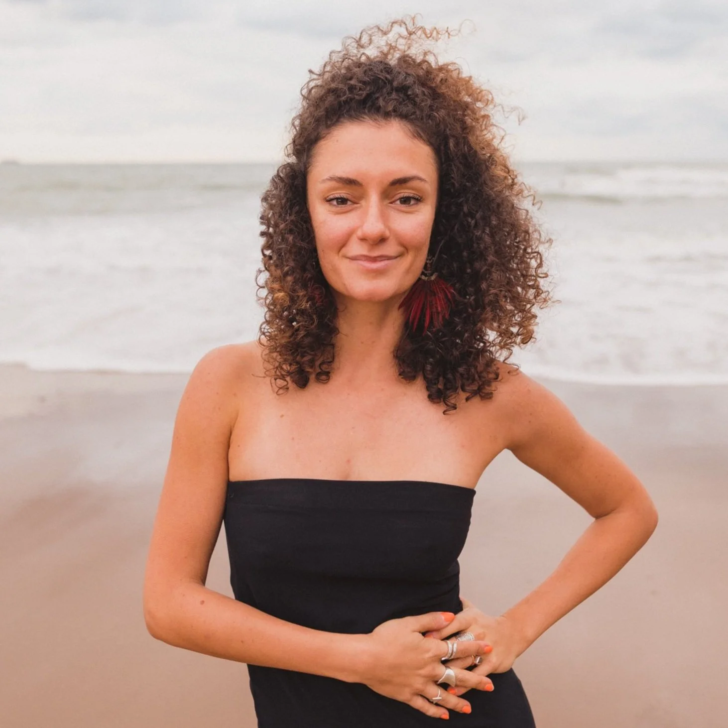 Breathwork Facilitator, Isik Tlabar stands at the beach, looking and smiling at the camera