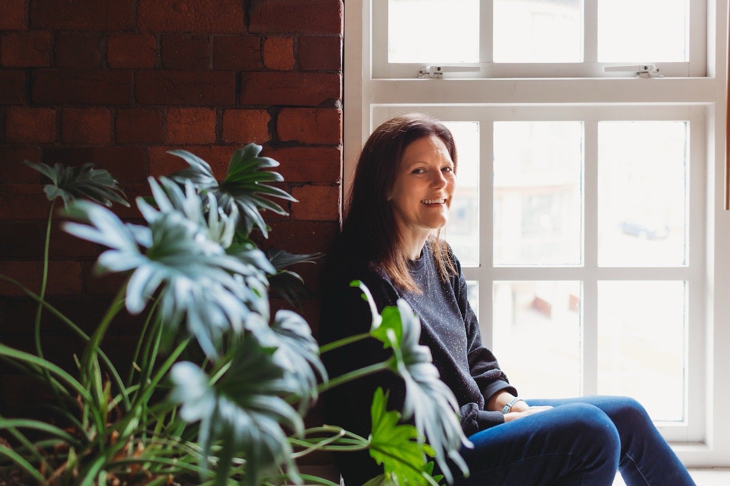 Architect Amanda Spence sat infant of large window with exposed brick wall and house plant