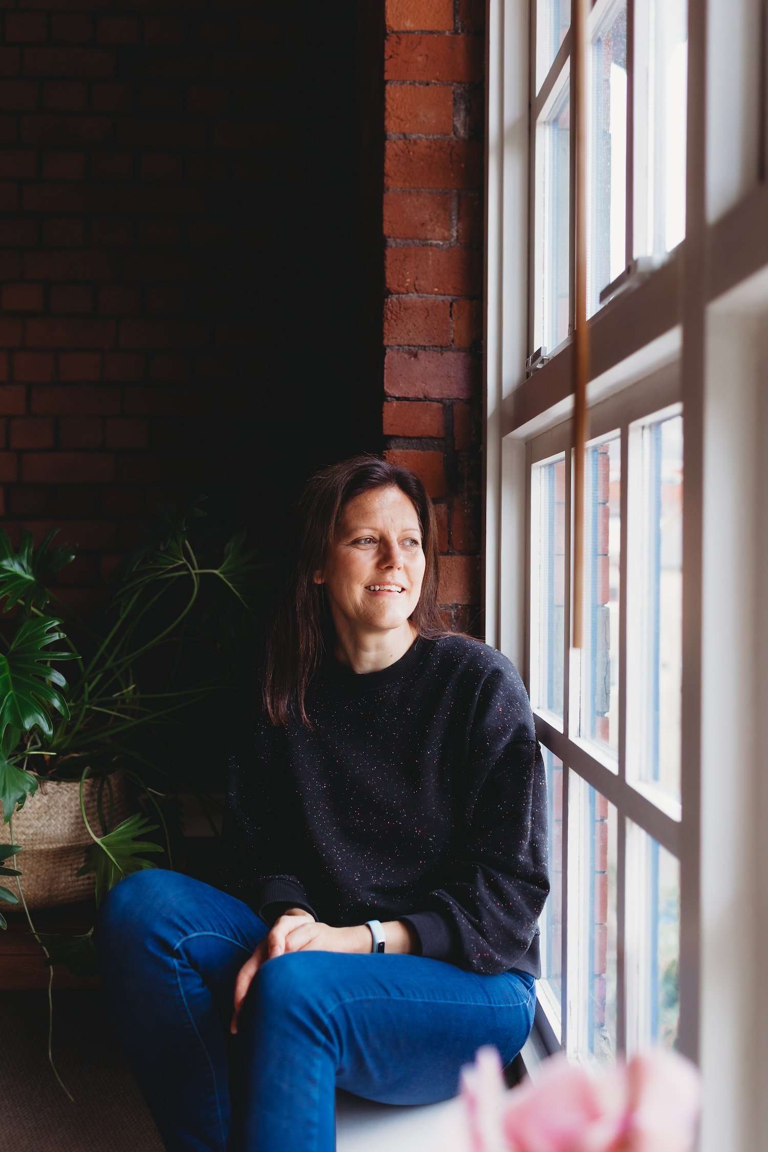 Architect Amanda Spence sat on window sill looking out of large window, exposed brick wall, large house plant in background