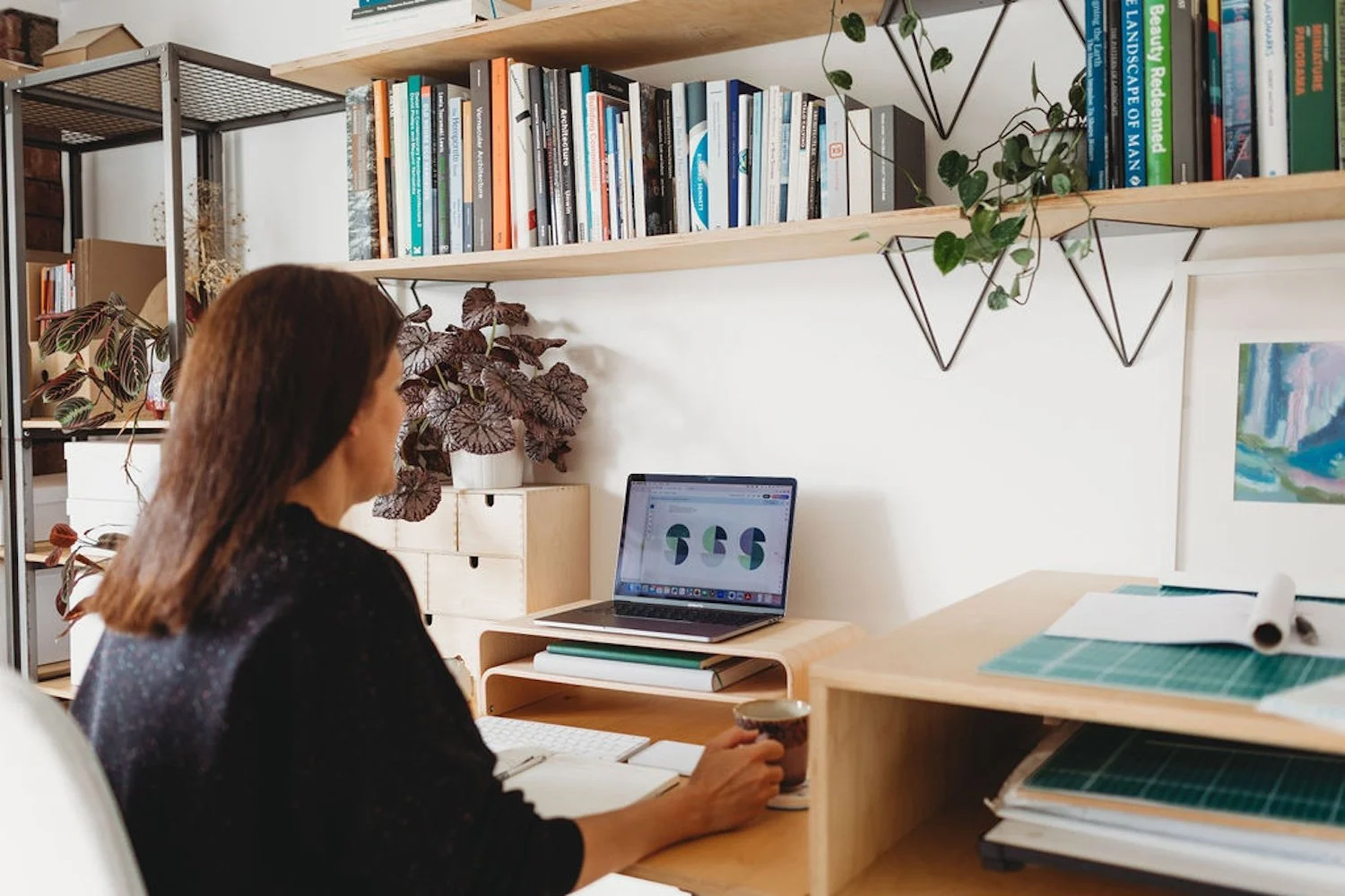 Architect Amanda Spence sat at desk in studio working on laptop with shelf of books and plants
