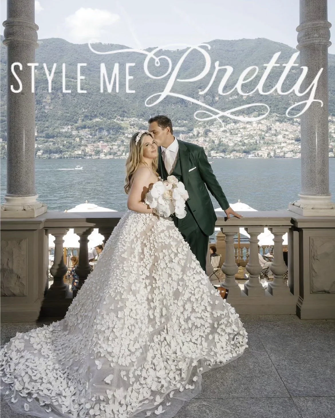 A bride and groom sharing a kiss on a balcony overlooking a body of water and mountains in the background.